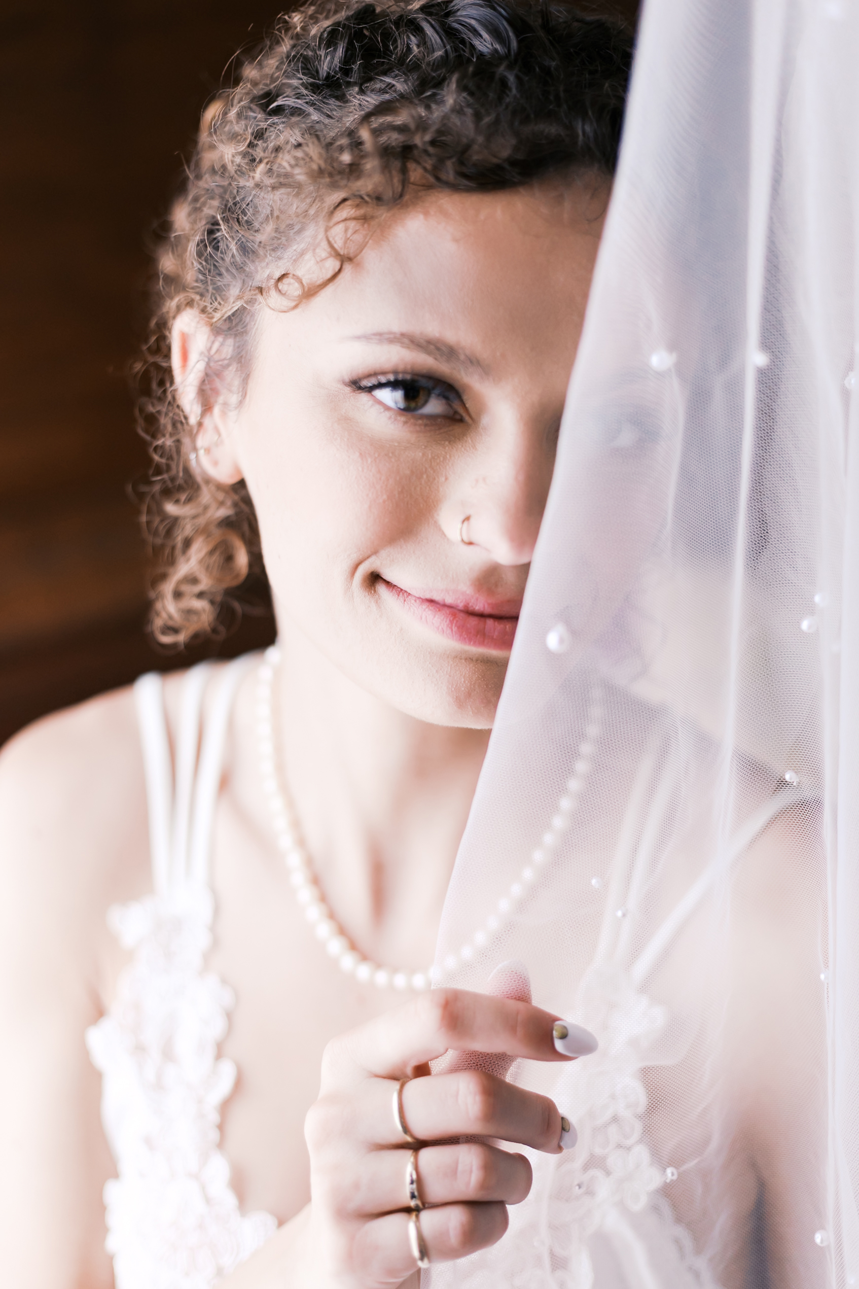 Close-up of bride’s face behind a dotted veil, showing her soft smile, pearl necklace, and delicate rings.