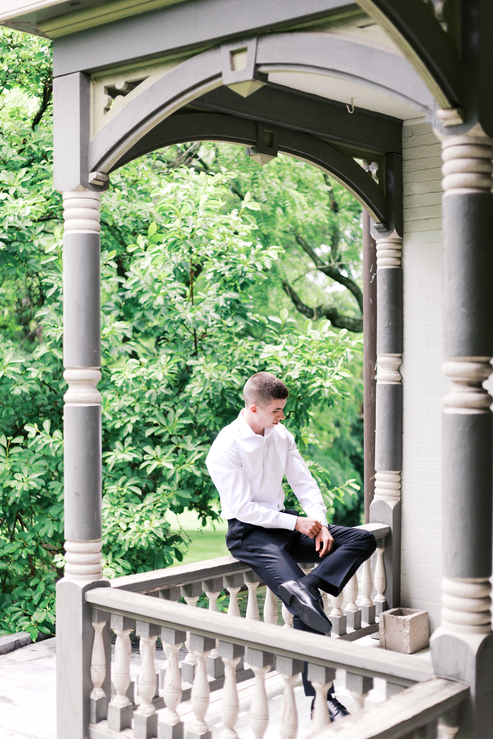 Groom in a white shirt and black slacks sits on the railing of a Victorian-style balcony surrounded by lush greenery.