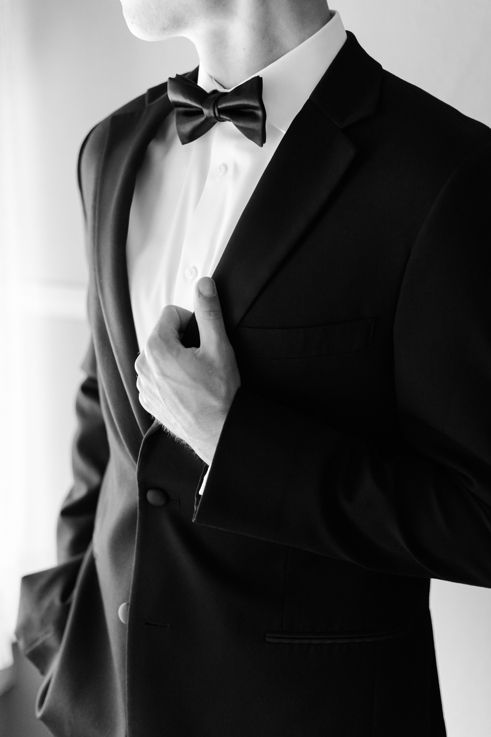 Monochrome close-up of a groom’s hand adjusting his suit jacket, showing crisp lapels and a black bow tie.