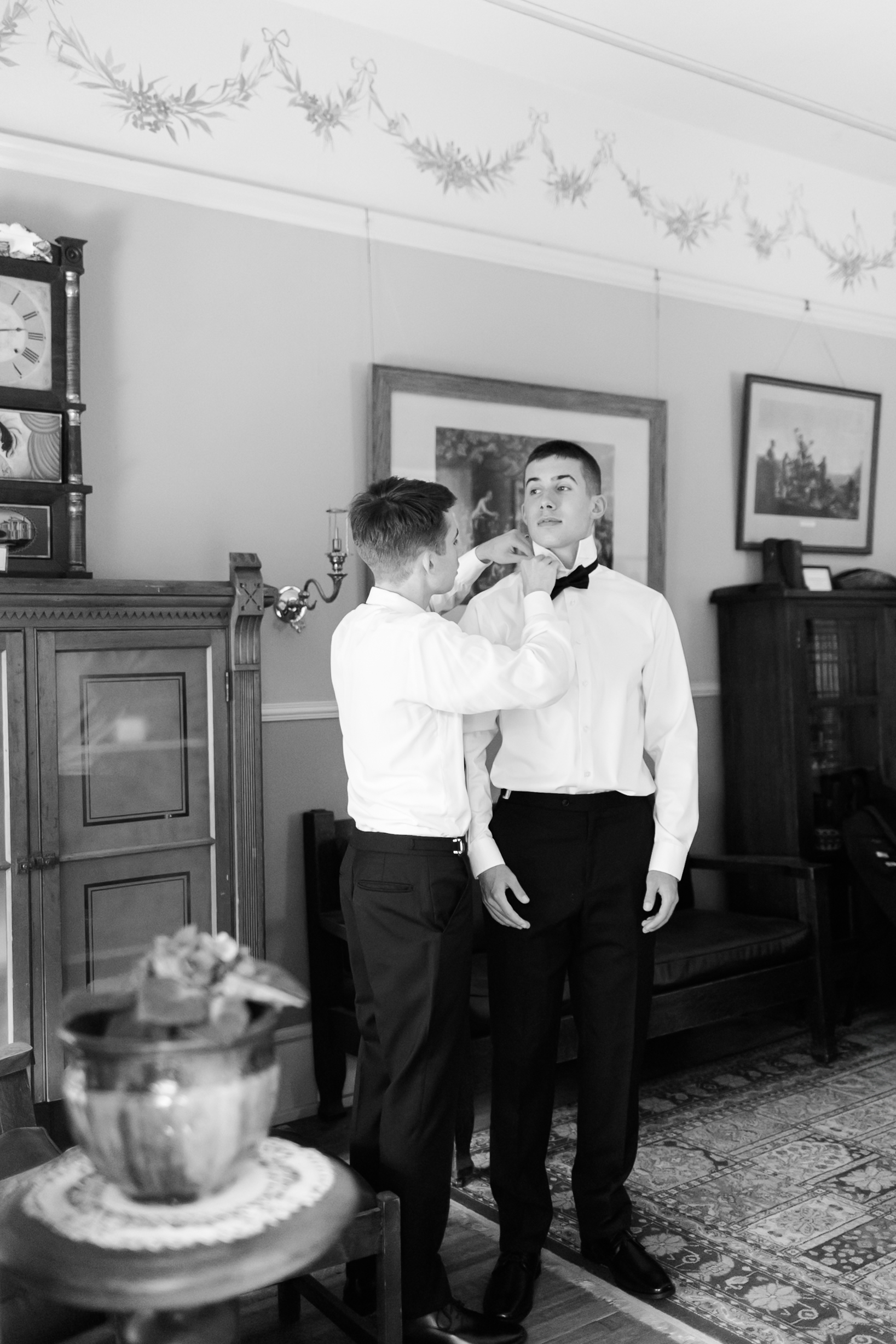 Black and white shot of two men in dress shirts, one adjusting the other’s bow tie in a traditional parlor setting.