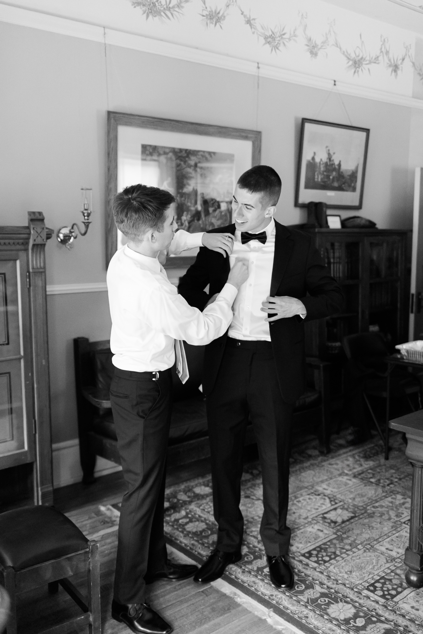 Two groomsmen smiling as one adjusts the other’s bow tie in a warmly lit, vintage-style room.