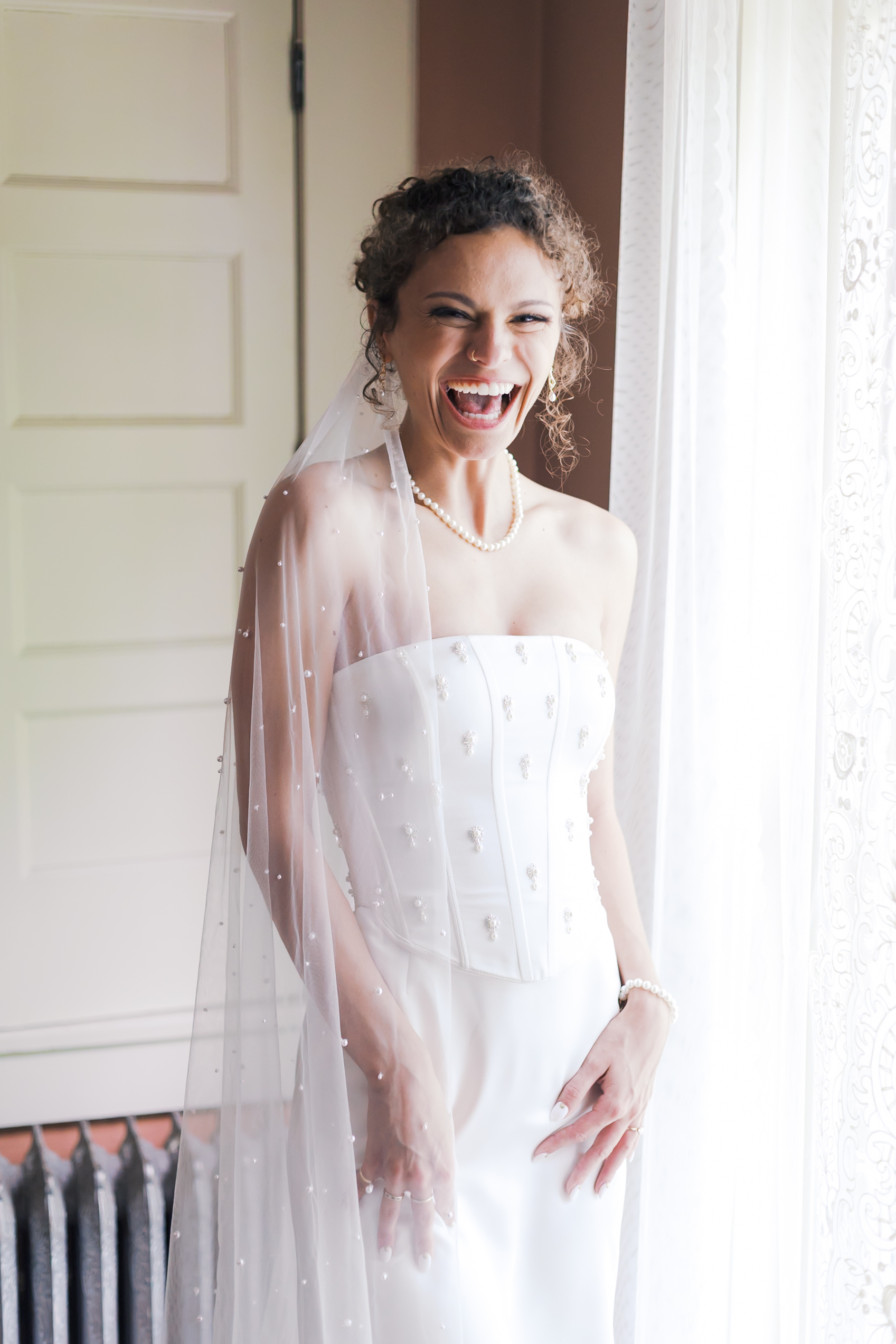 Bride in a pearl-detailed gown and veil laughing joyfully beside a sunlit window with lace curtains.