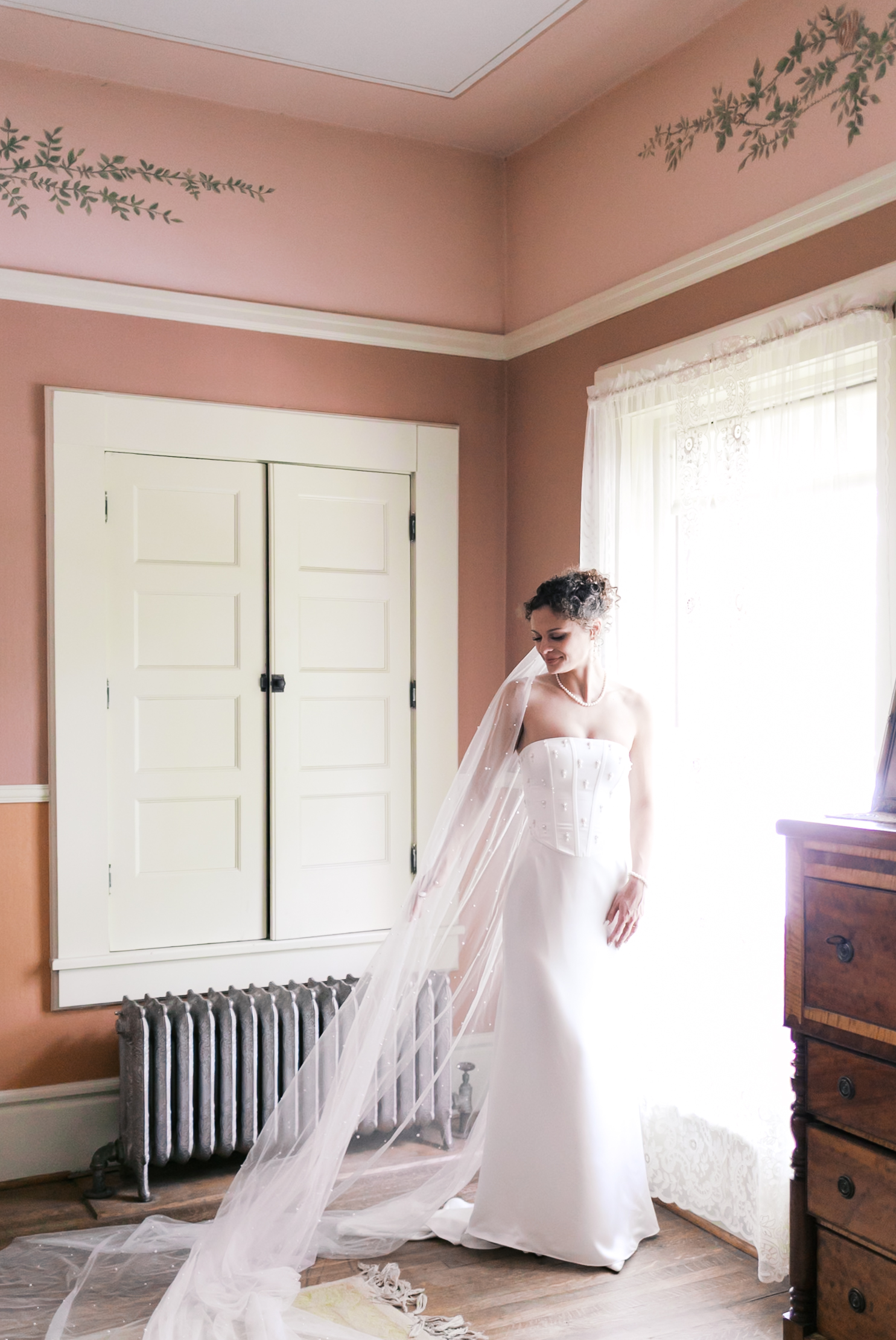 Bride standing gracefully in a vintage-style room, holding the train of her long dotted veil near a lace-draped window.