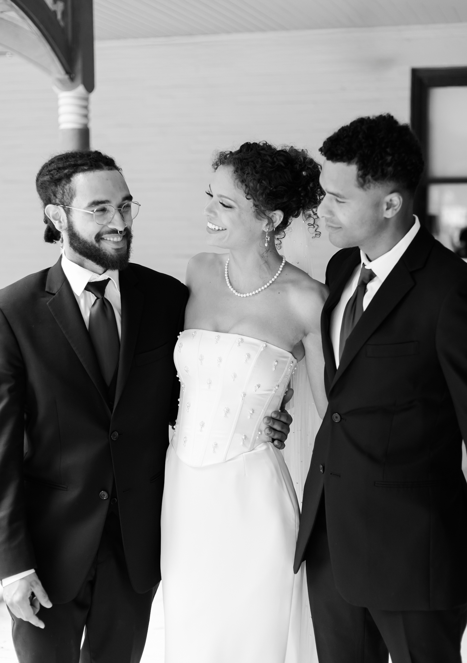 Black and white portrait of a bride smiling at two men in suits, standing together on a porch with vintage architecture.