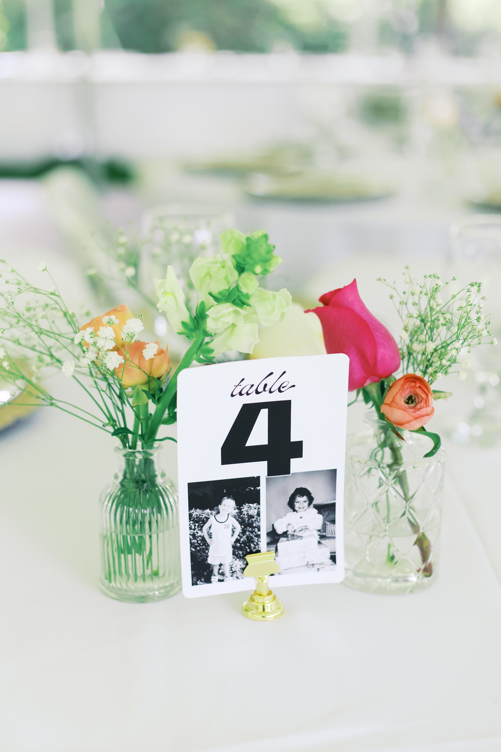 Table number card with black-and-white childhood photos, surrounded by small glass vases of colorful flowers.