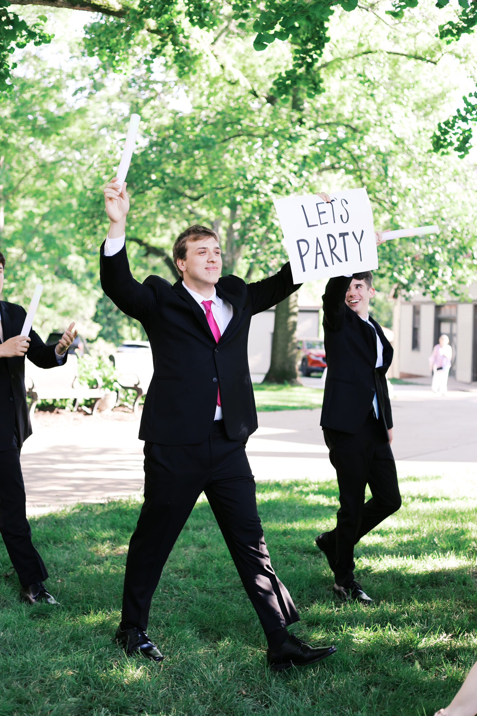 Groomsman in black suit and pink tie walks across grass holding a “LET’S PARTY” sign, smiling with fellow groomsmen.