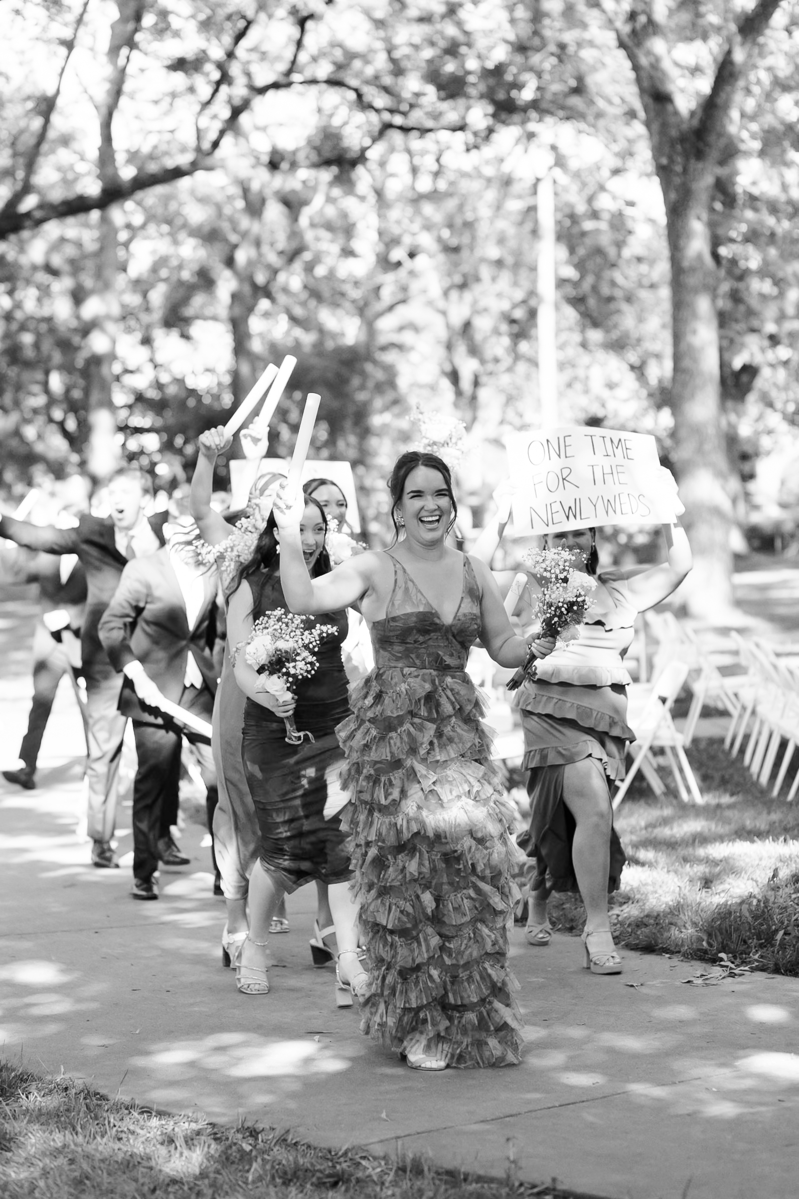 Black and white image of a smiling and cheering woman followed by a small group of people, one person holding a sign that says "one time for the newlyweds"