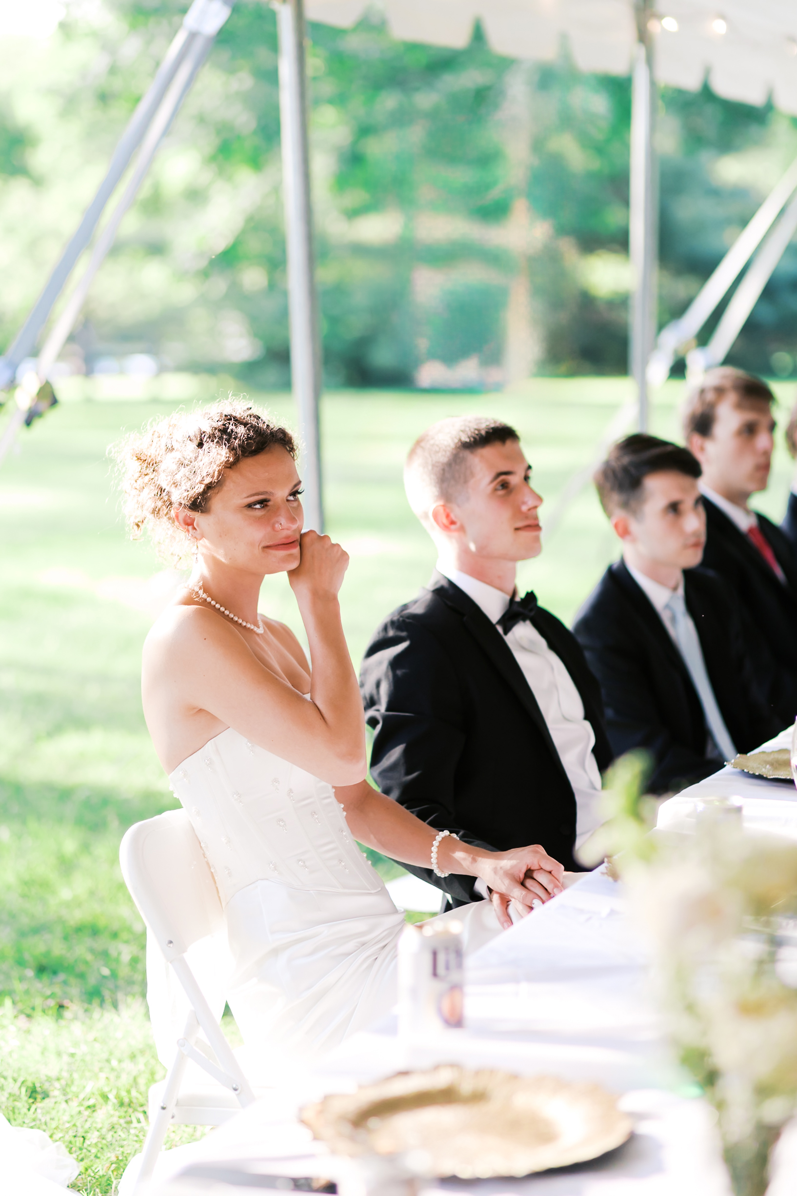 Bride holding her groom's hand as they sit watching toasts.