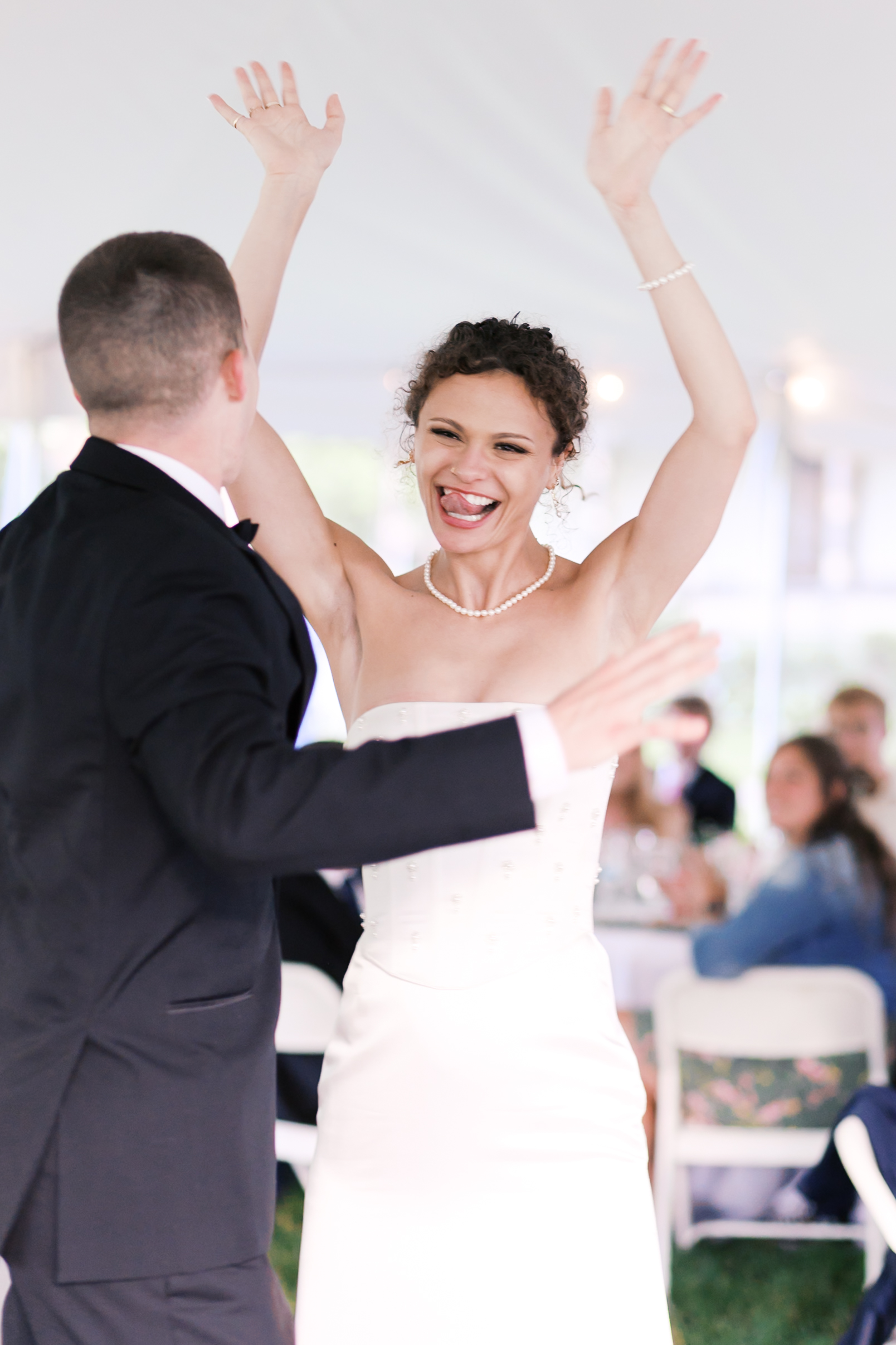 Bride with her tongue out and smiling as she raises her hands in celebration with the groom dancing in the foreground.