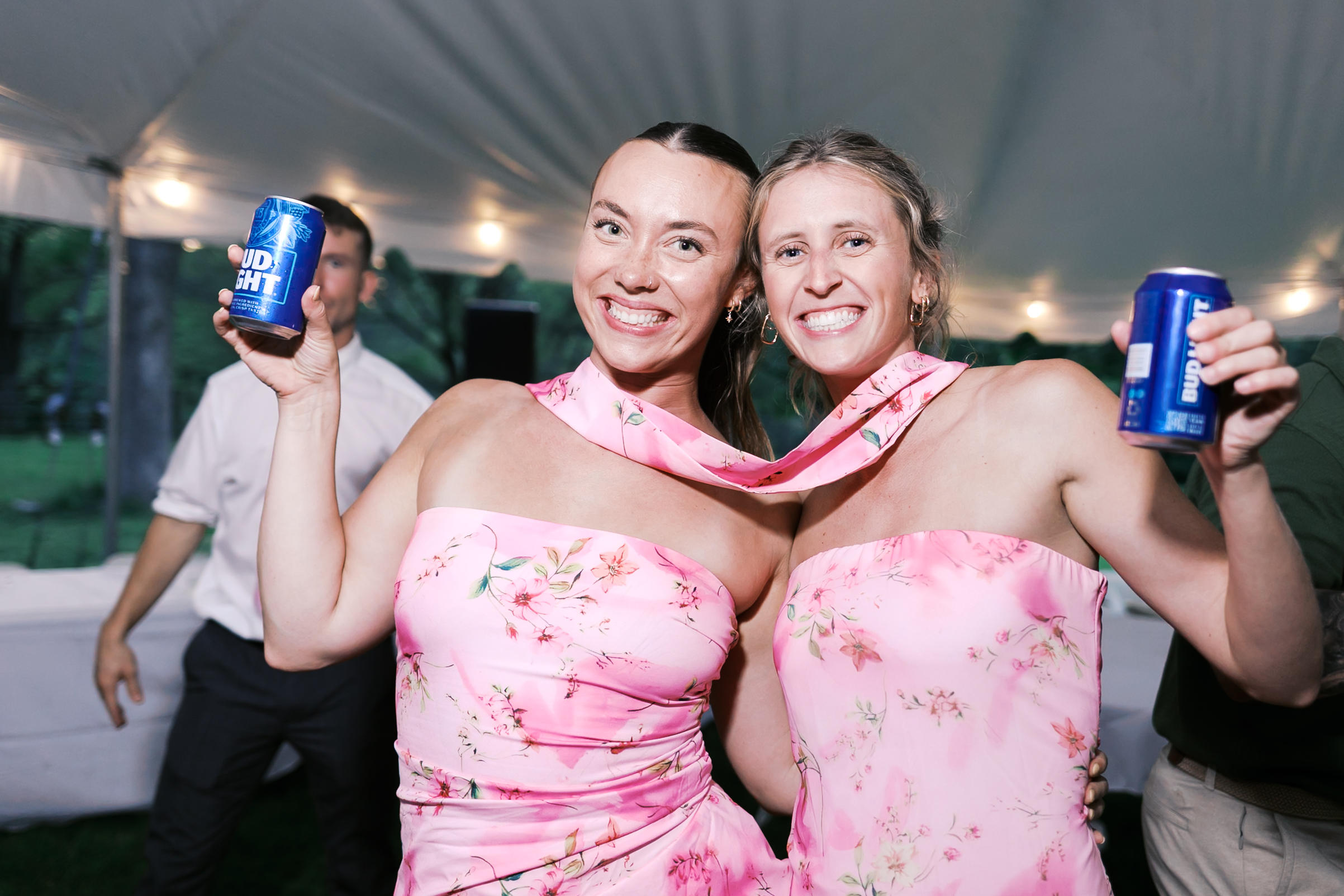 Wedding guest in pink floral dresses smile at the camera while holding blue Bud Light cans.