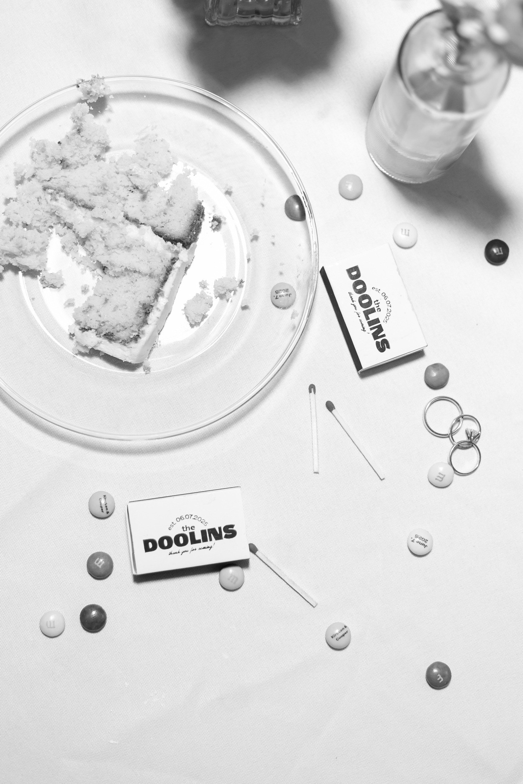 A black and white flat lay image of eaten cake, custom m&m's, wedding rings, and custom matchboxes that say the Doolins.