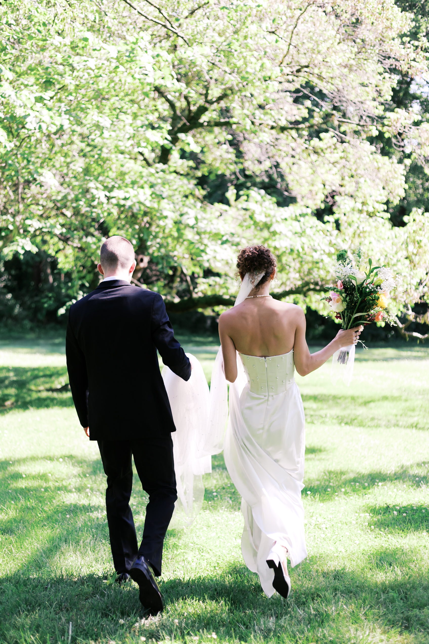 Bride in a white gown and groom in a black suit walk away hand in hand across a bright, grassy field.