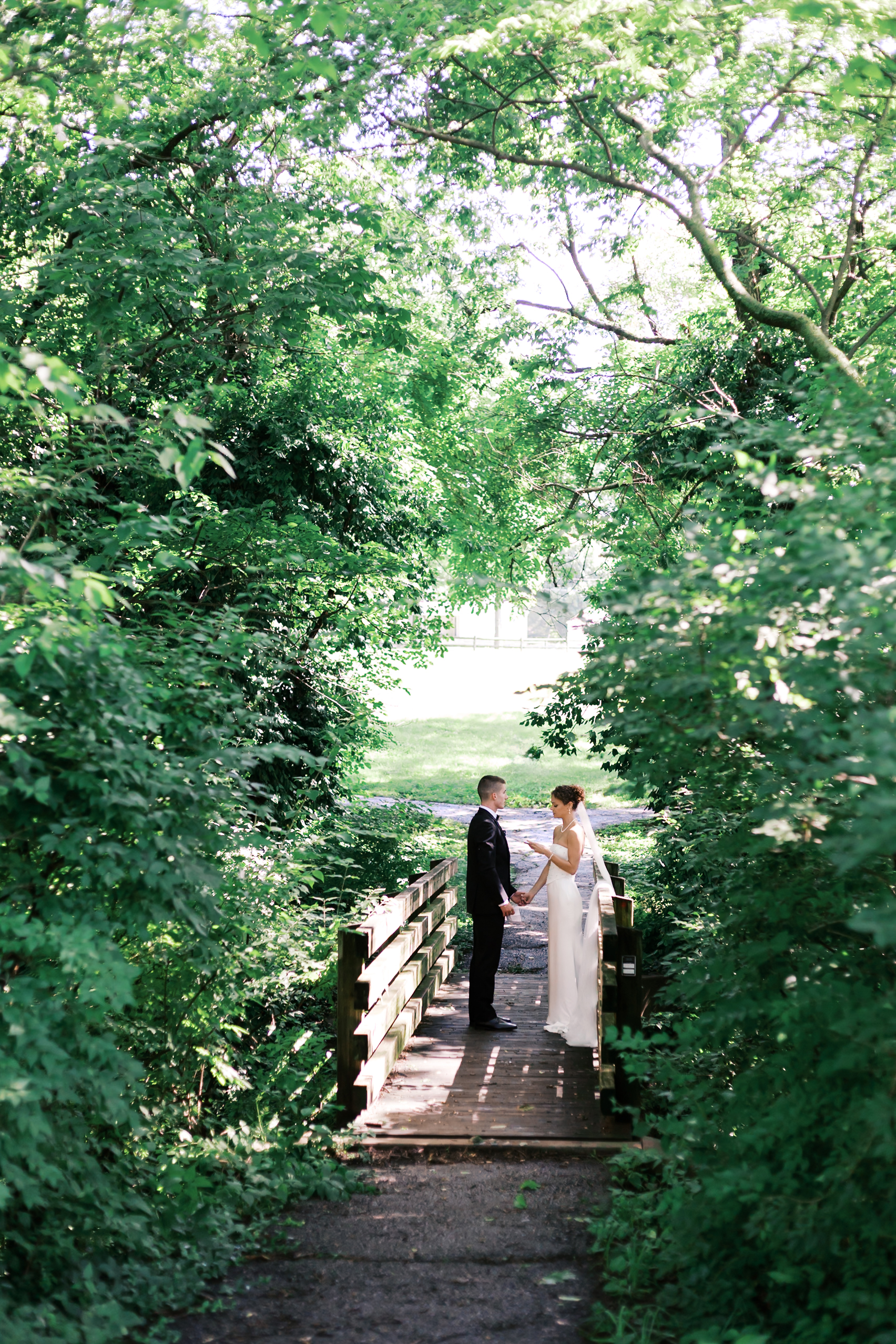 Bride and groom stand face to face holding hands on a small wooden bridge surrounded by lush green trees.