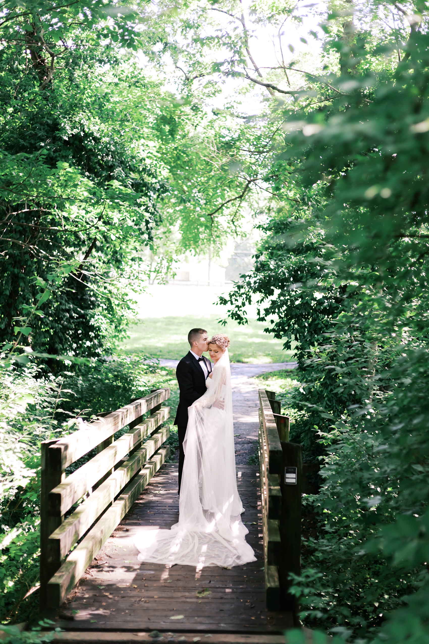 Groom gently kisses bride’s forehead as they stand embraced on a wooden bridge in a forest clearing.