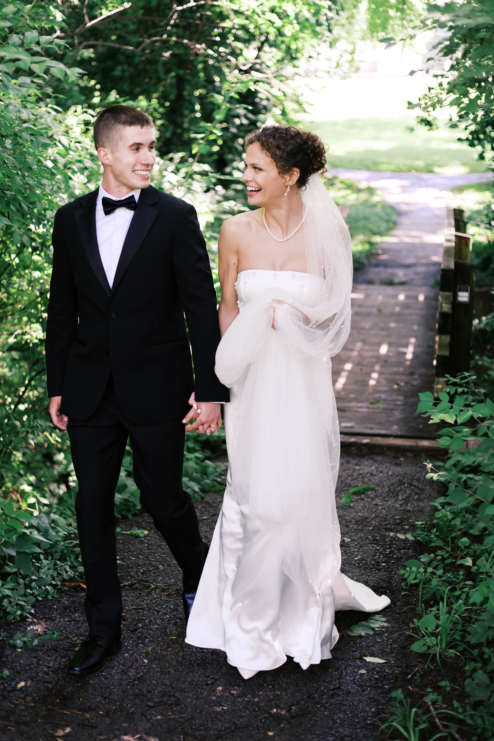 Smiling bride and groom walk together on a tree-lined path, holding hands near a rustic wooden bridge.