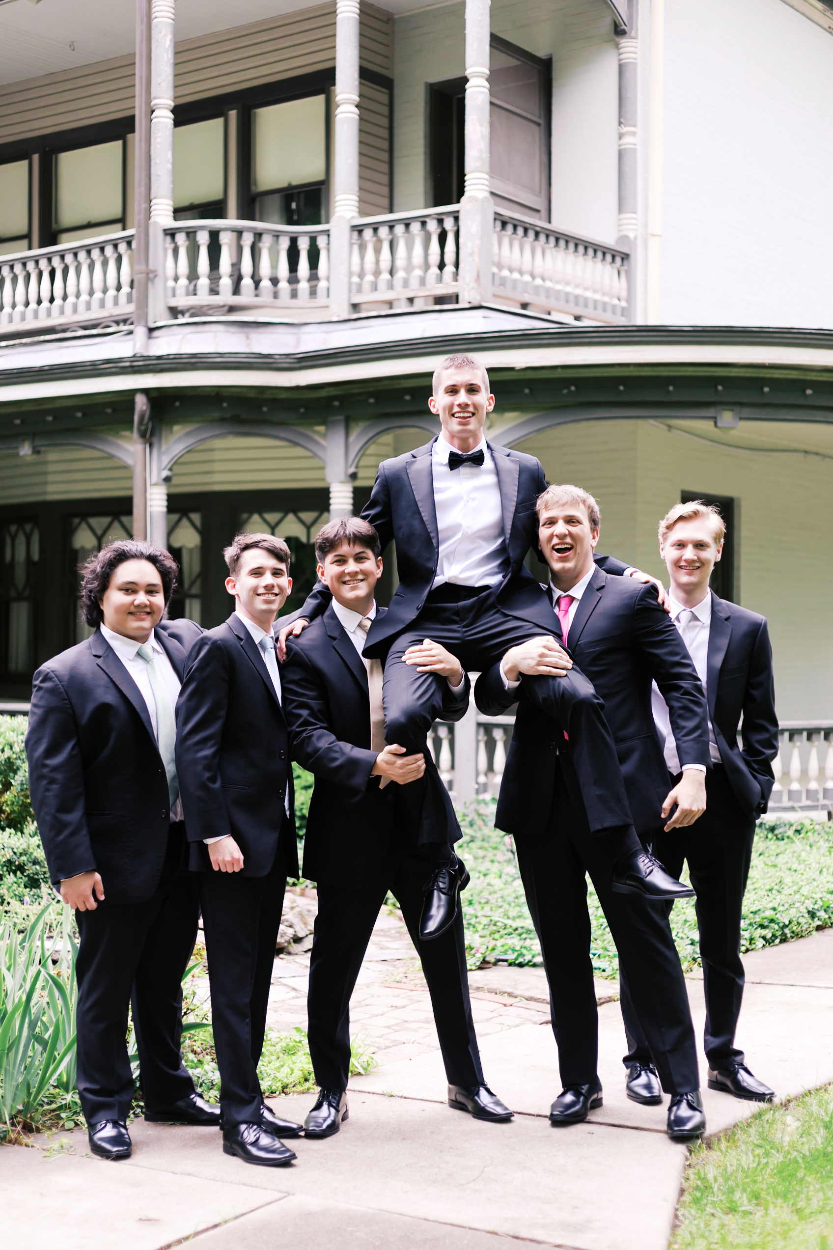 Groomsmen lift groom onto their shoulders in a playful group photo outside a historic building.
