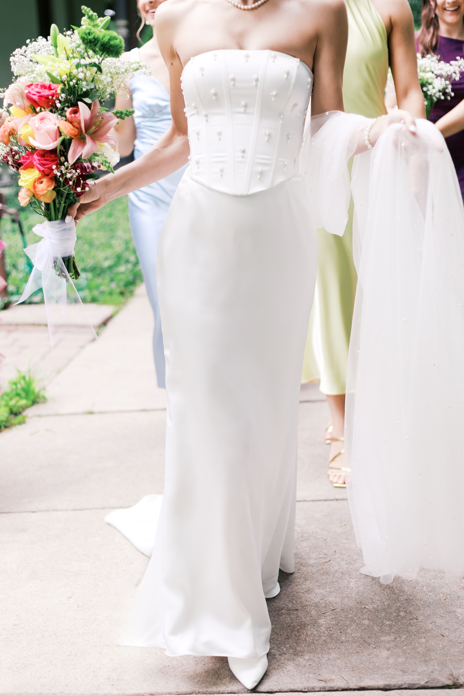 Detail of bride’s pearl-detailed gown and colorful bouquet as she walks forward with her bridal party.