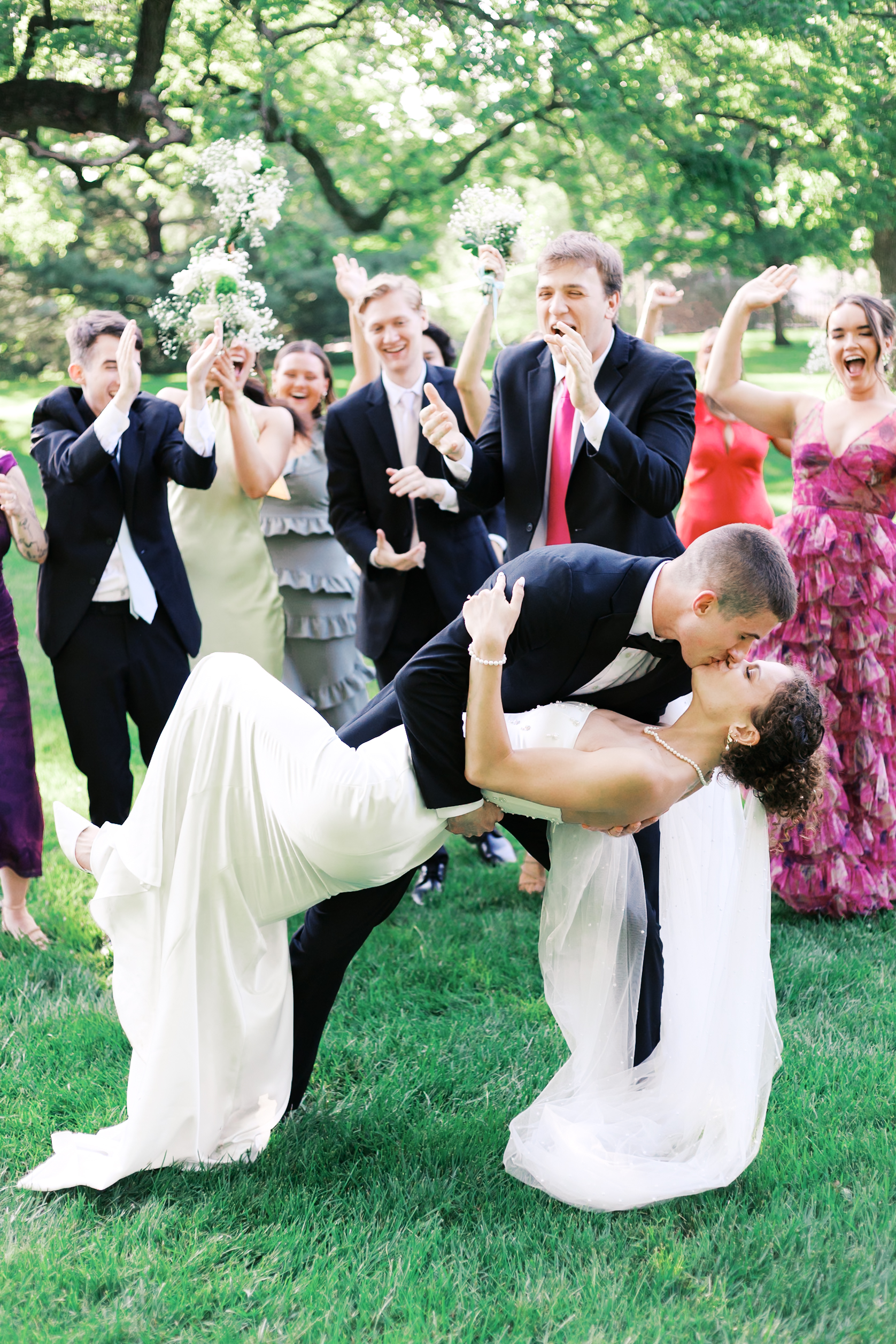 Groom dipping bride and kissing her as bridal party in colorful dresses and tuxedos look on and cheer.