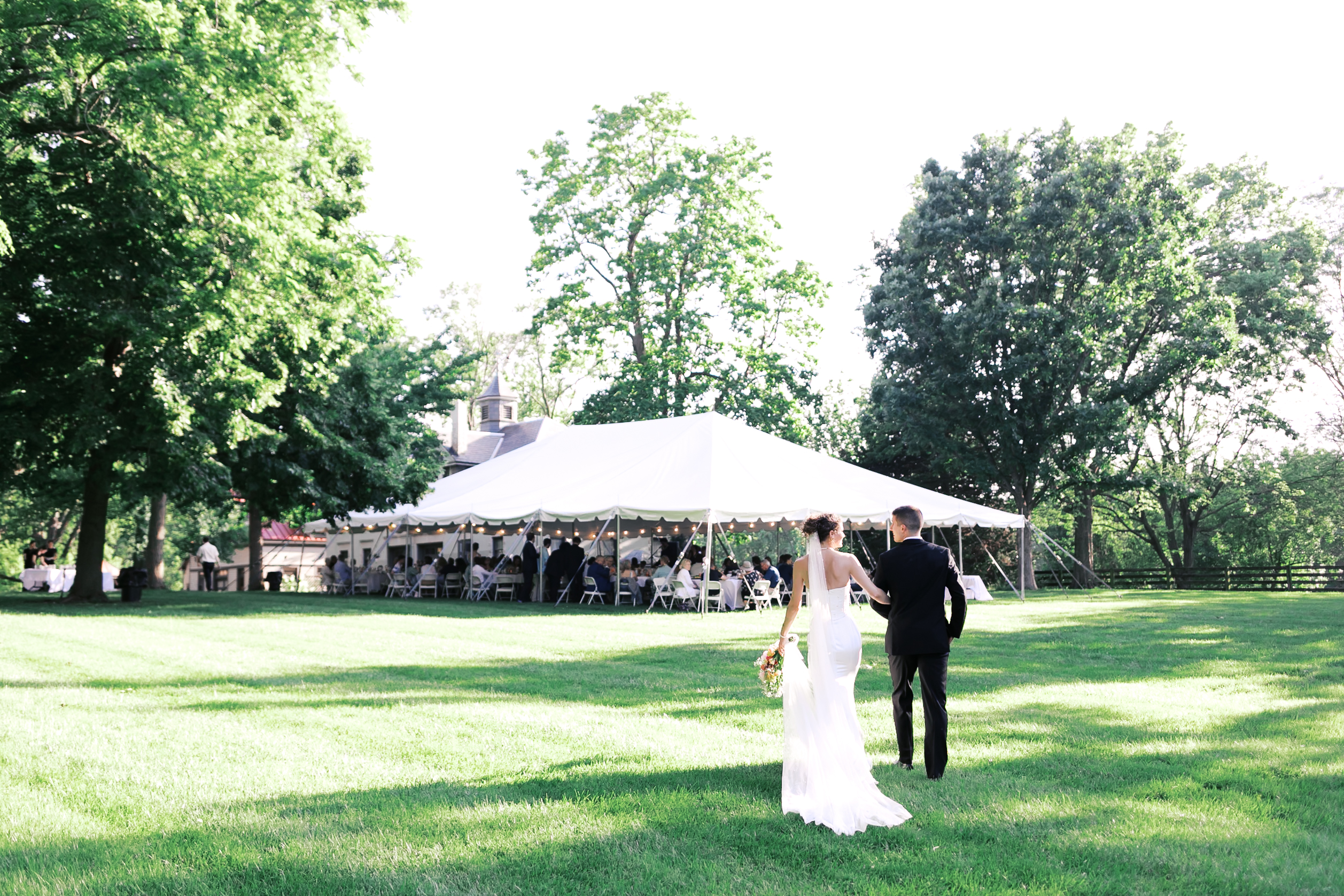 Bride looking at groom as they walk across a grass covered lawn toward a tented receptionf illed with guests.