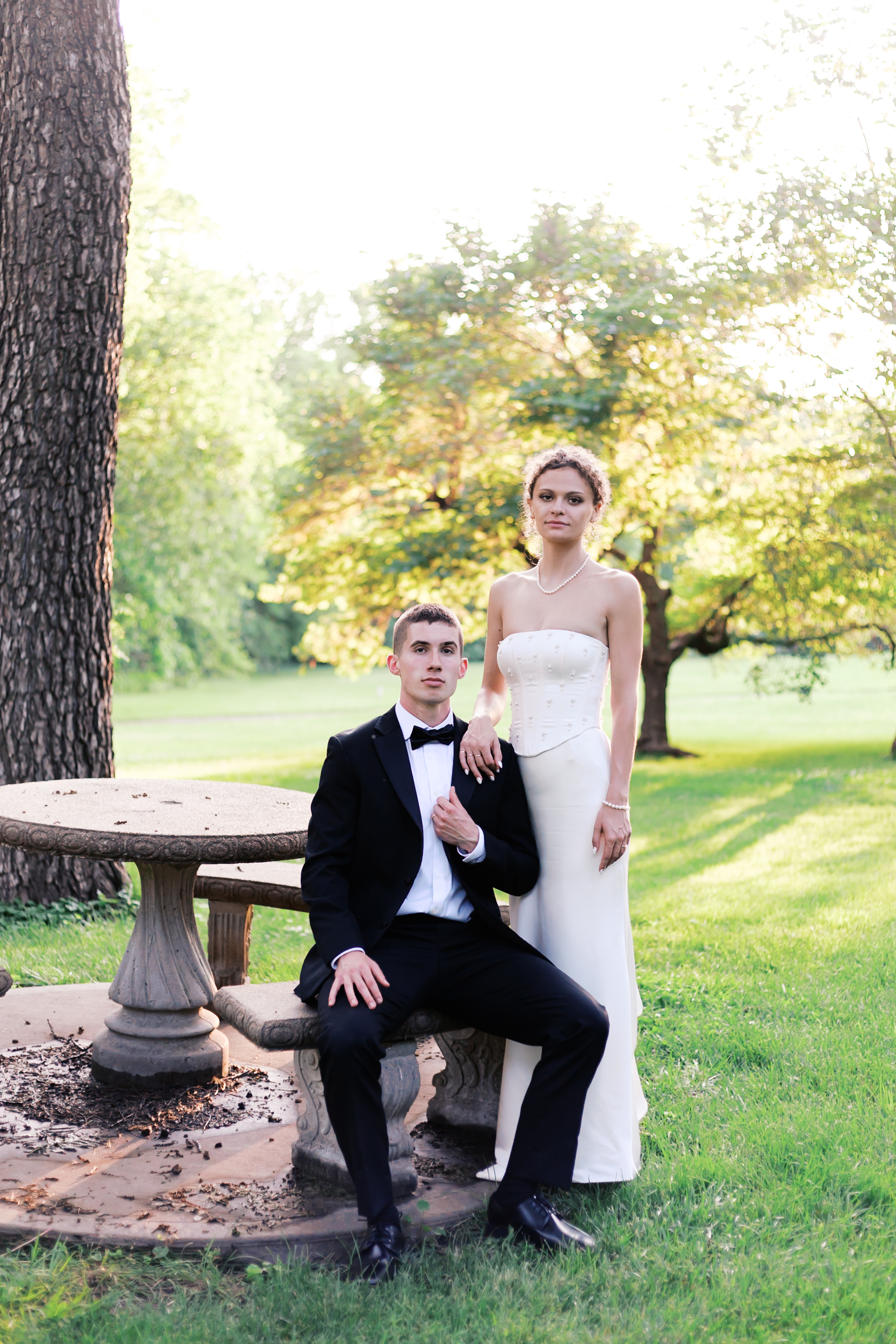 Bride stands beside groom who is seated at a rustic stone picnic table in a serene park setting during golden hour.