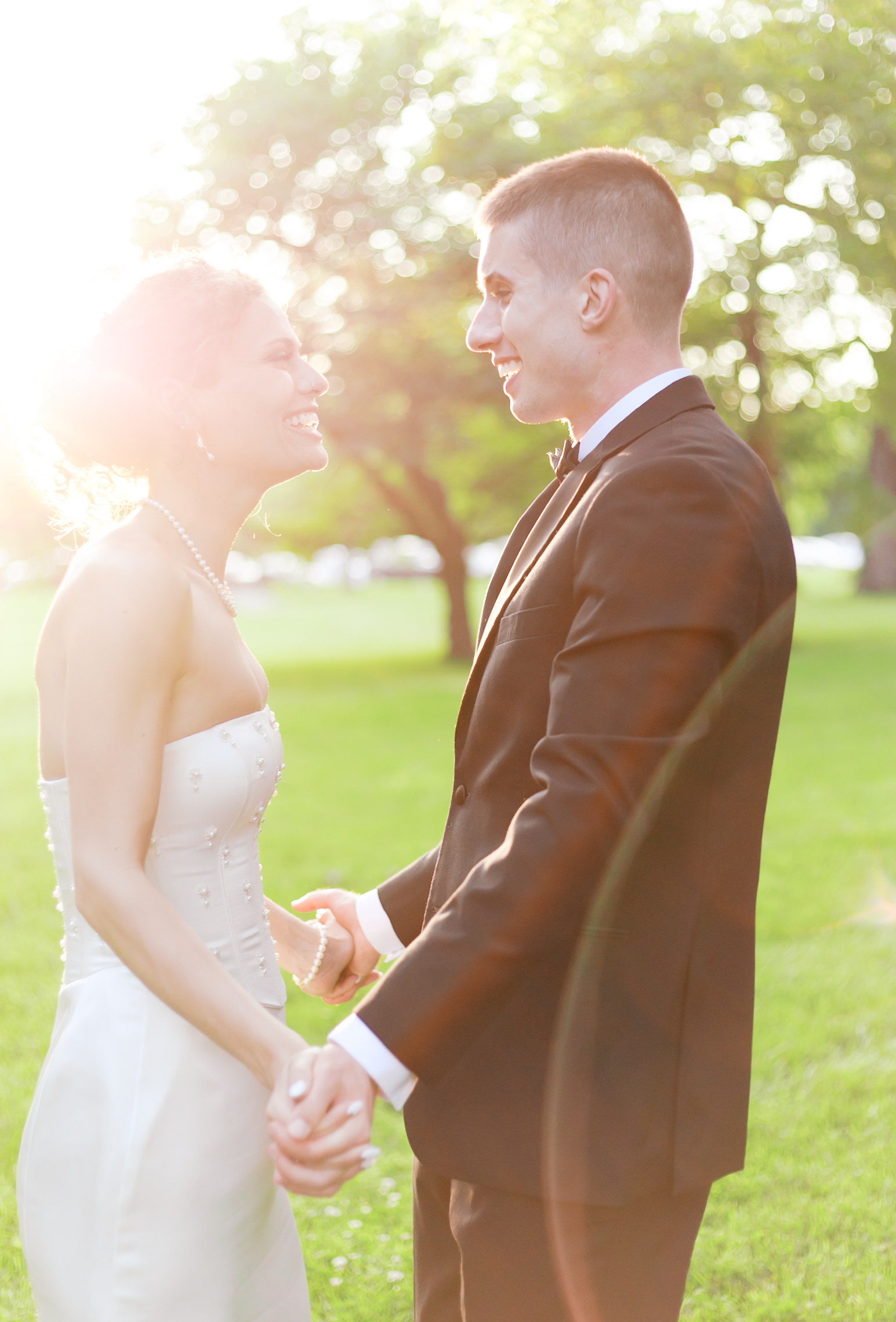 Bride and groom facing each other and holding hands in a sunny, backlit green space.