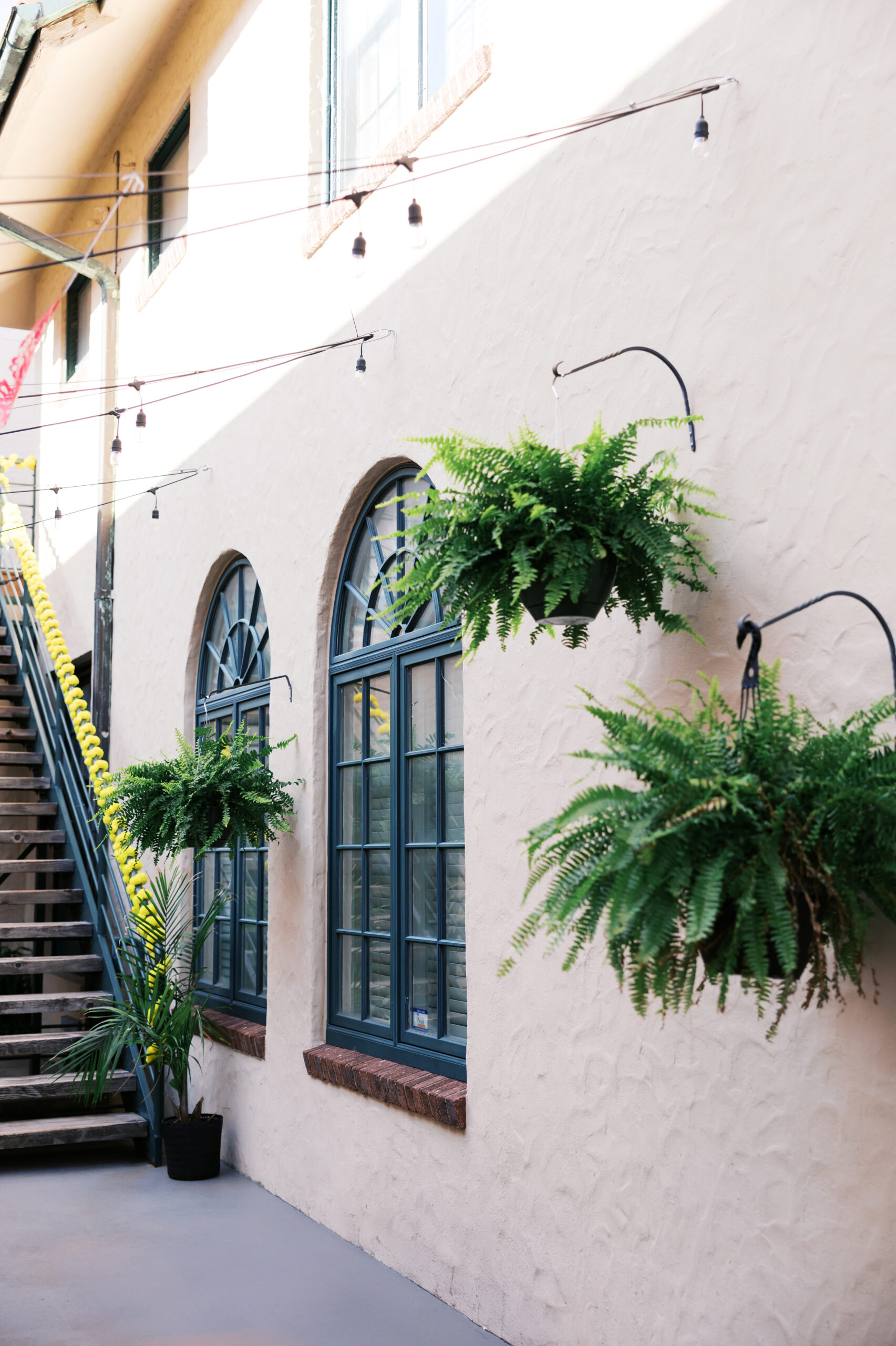 Stucco courtyard wall with arched windows, hanging ferns, and string lights at a Spanish-style event venue.