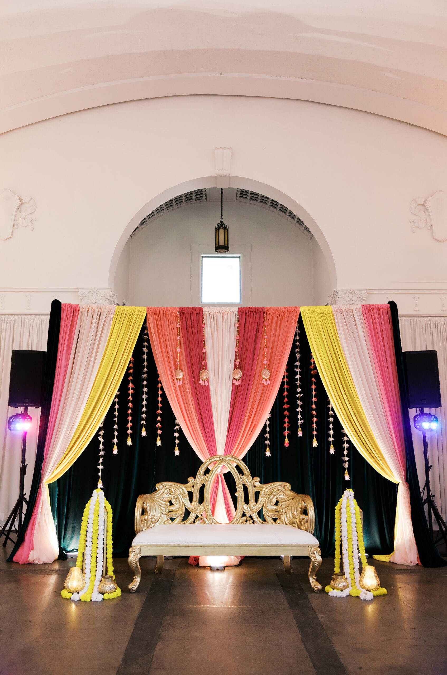 Indian wedding Jaggo stage with pink and yellow draping, floral garlands, and an ornate gold loveseat.