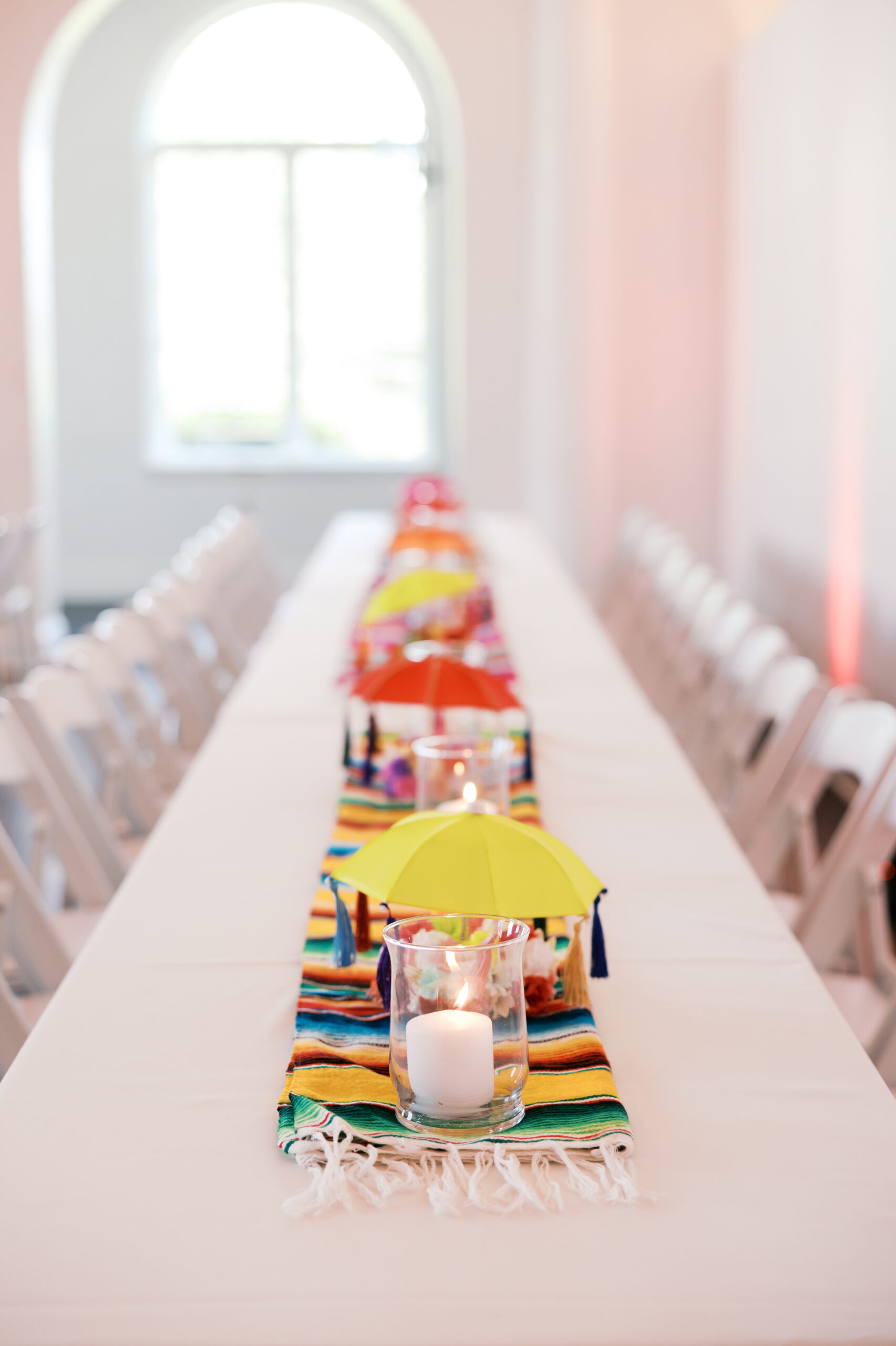 Colorful wedding centerpiece with a mini umbrella, candle, and striped runner on a long reception table.