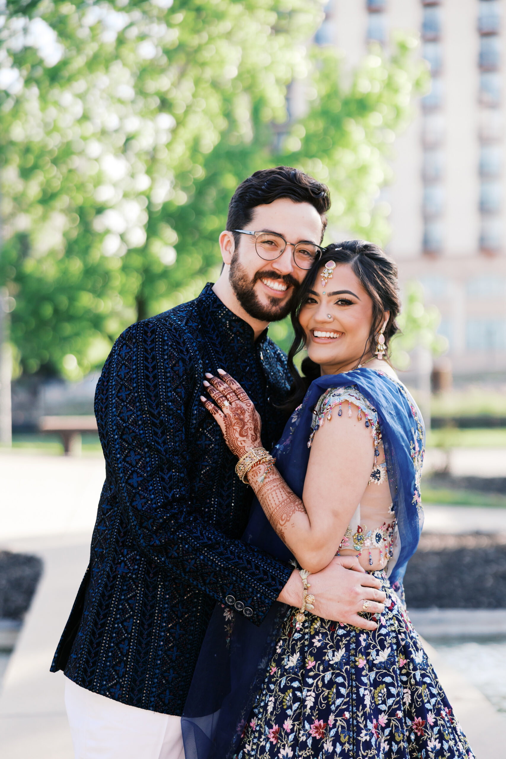 Couple embracing and smiling outdoors in traditional Indian wedding attire.