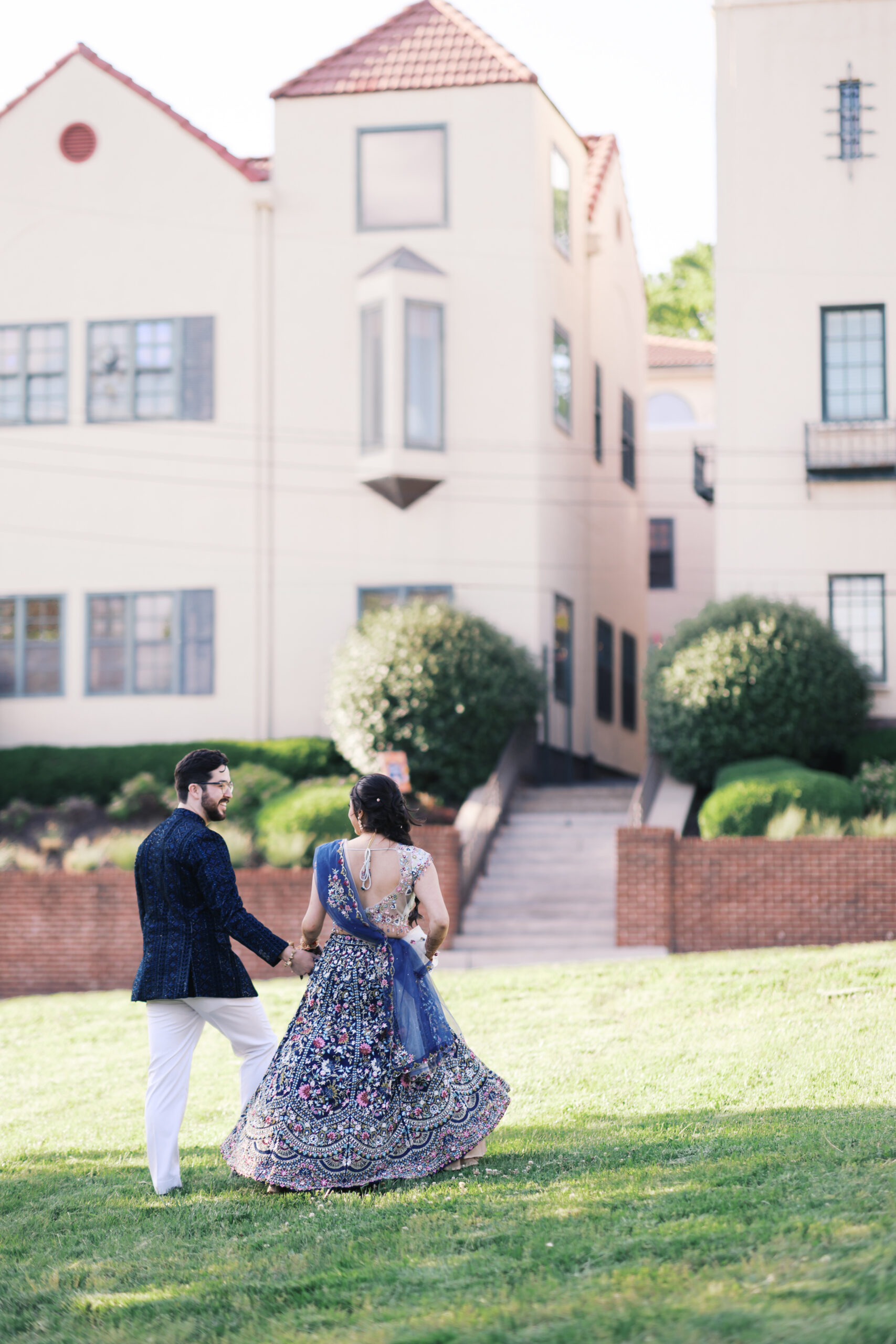 Couple holding hands on lawn in front of Spanish-style wedding venue.