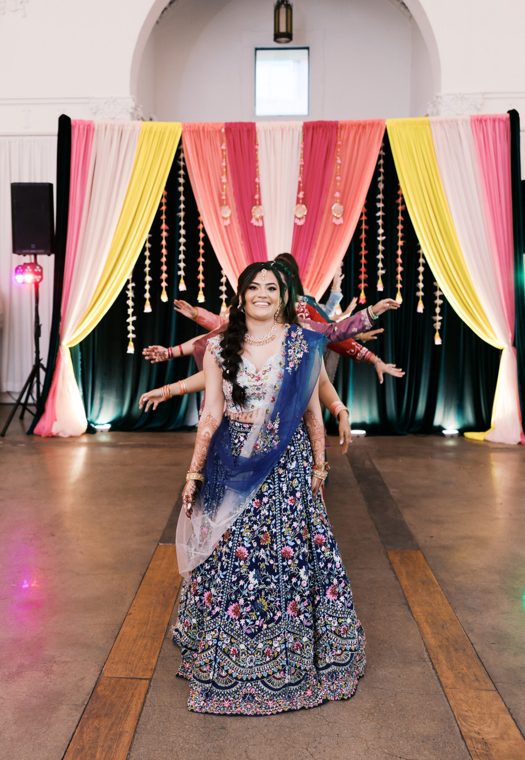 Bride dancing in floral lehenga in front of pink and yellow Jaggo backdrop.