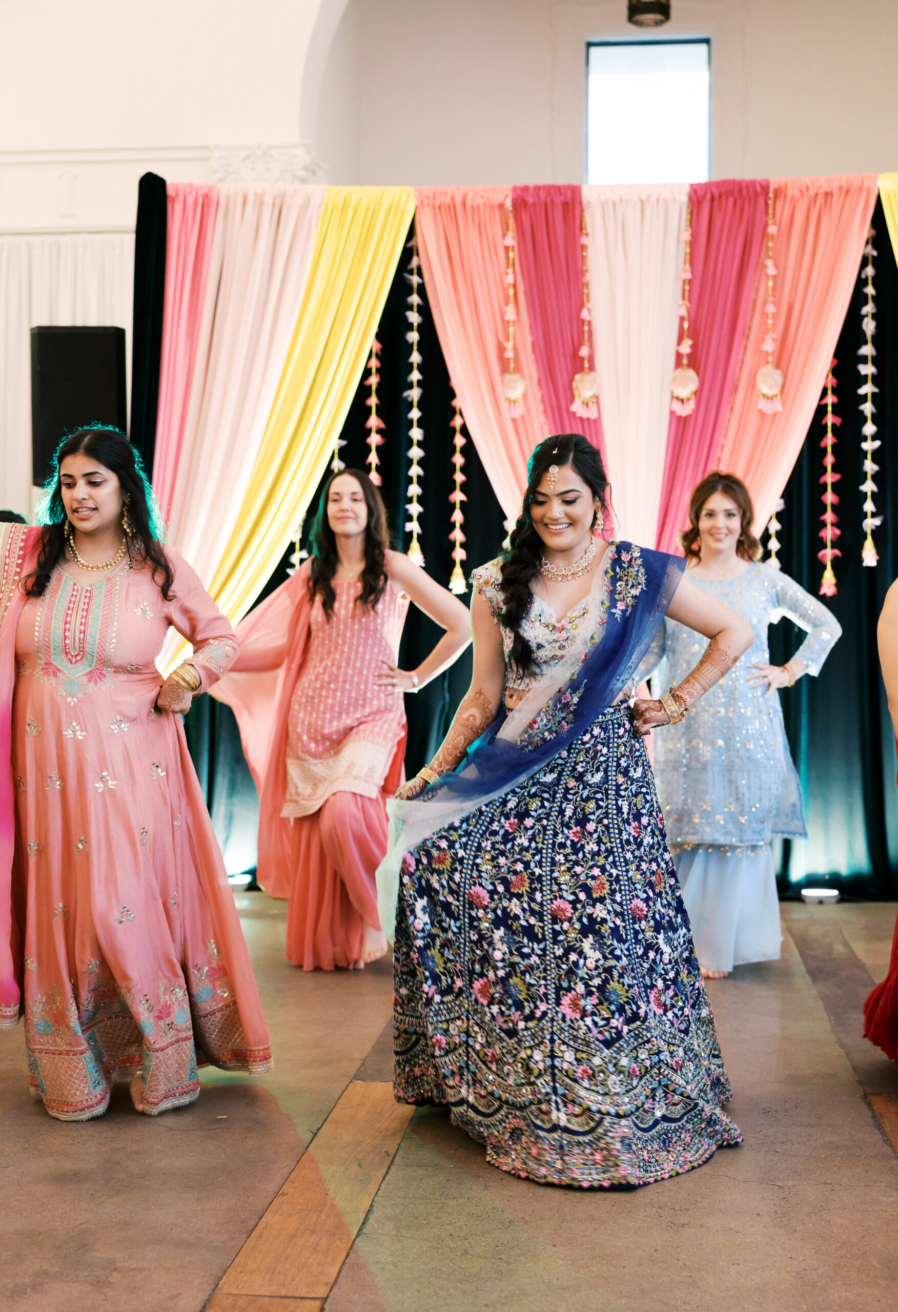 Bridesmaids dancing in pastel outfits during indoor South Asian wedding event.
