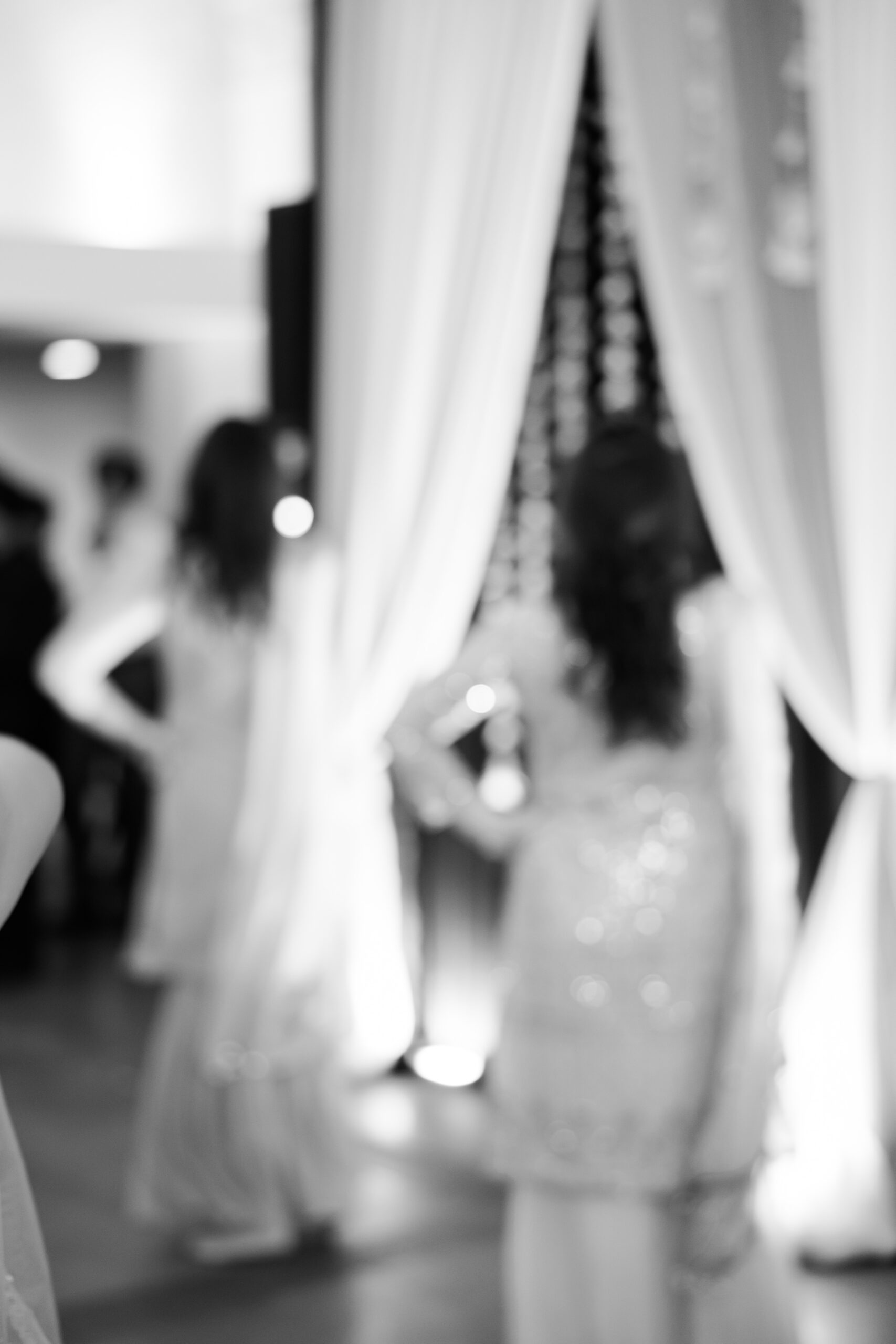 Soft black and white photo of guests dancing behind draped wedding backdrop.