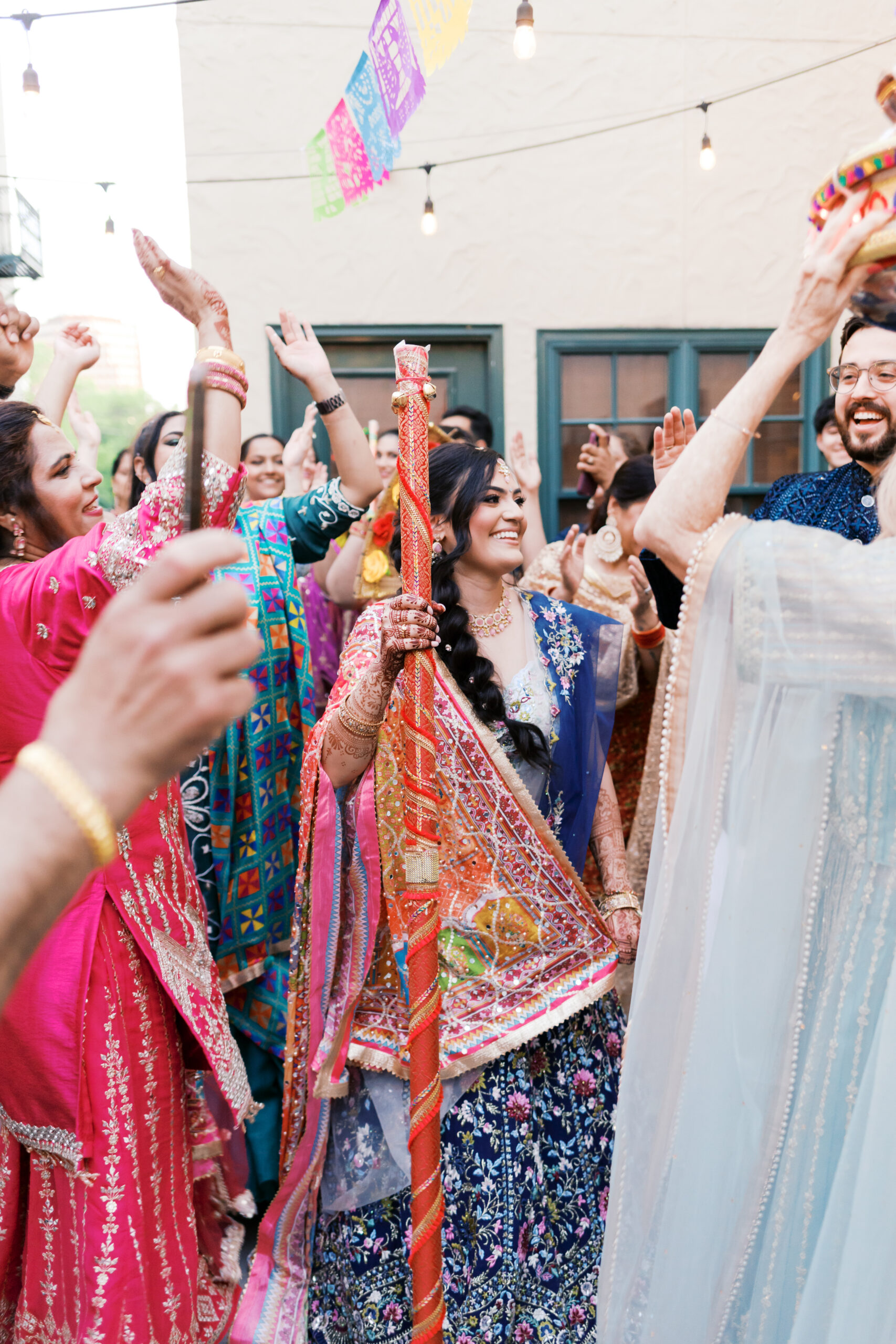 Bride holding decorated Jaggo stick while dancing with family outdoors.