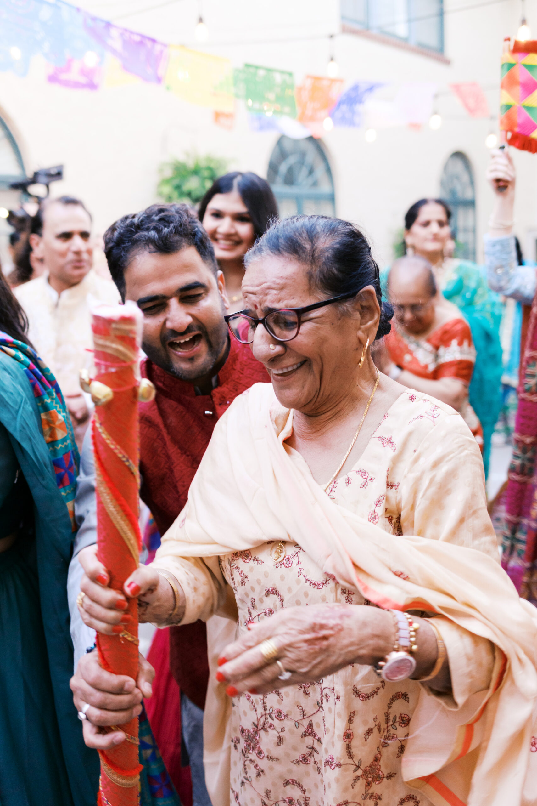 Older guest dancing with decorated stick during vibrant South Asian wedding event.