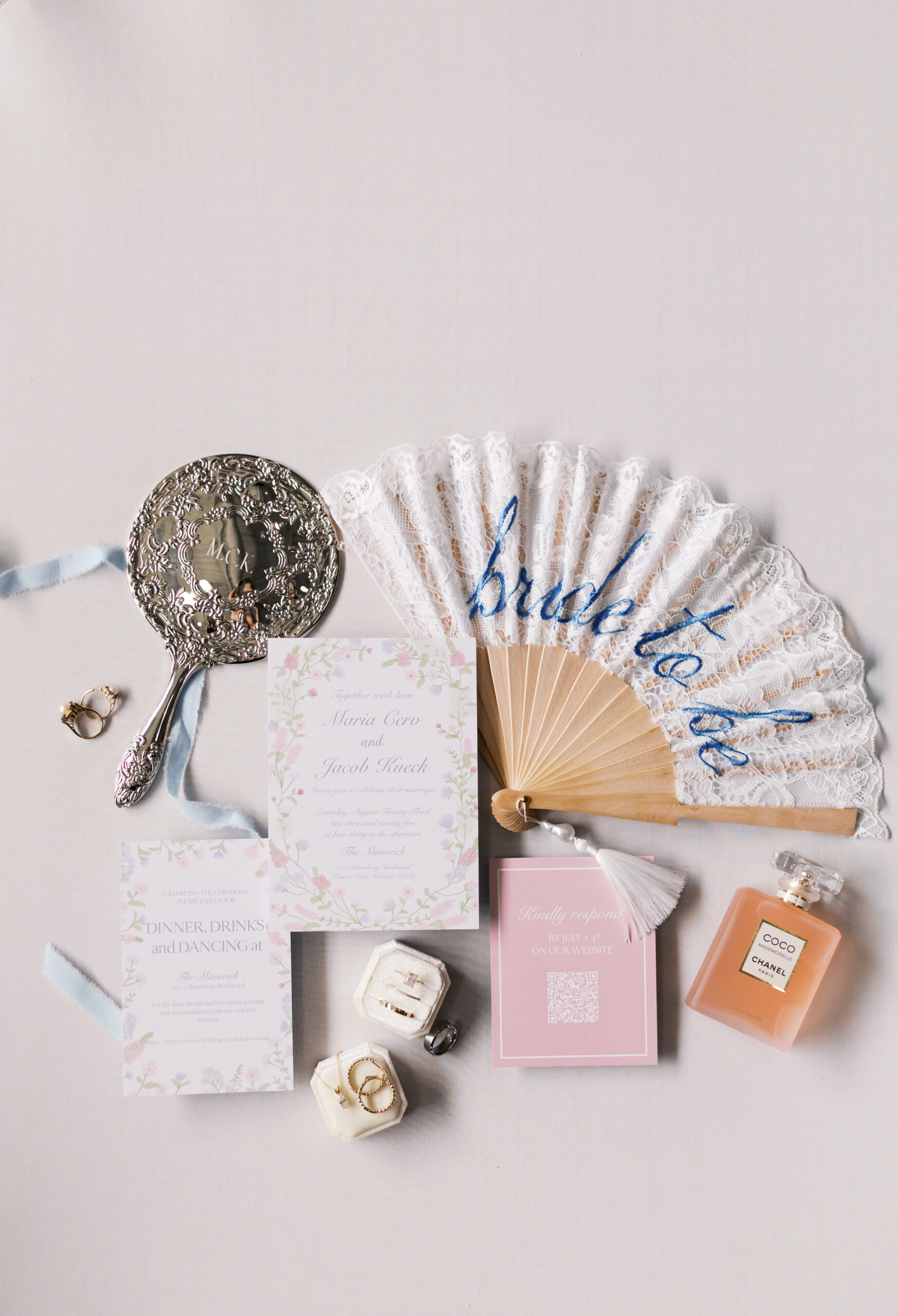 Wedding flat lay with invitation suite, lace “bride to be” fan, perfume, rings, and engraved hand mirror on a white background.
