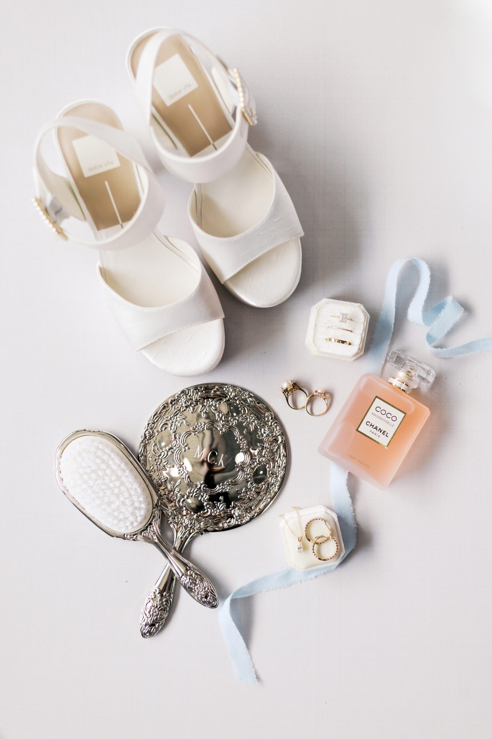 Bridal accessories flat lay featuring white heels, perfume bottle, wedding rings, and engraved silver mirror on a light background.
