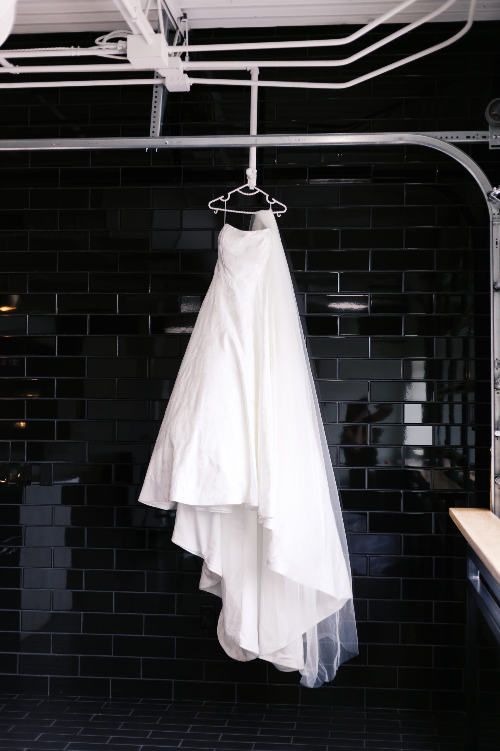 White strapless wedding dress hanging on a hanger against a black tiled wall in a modern bridal suite.
