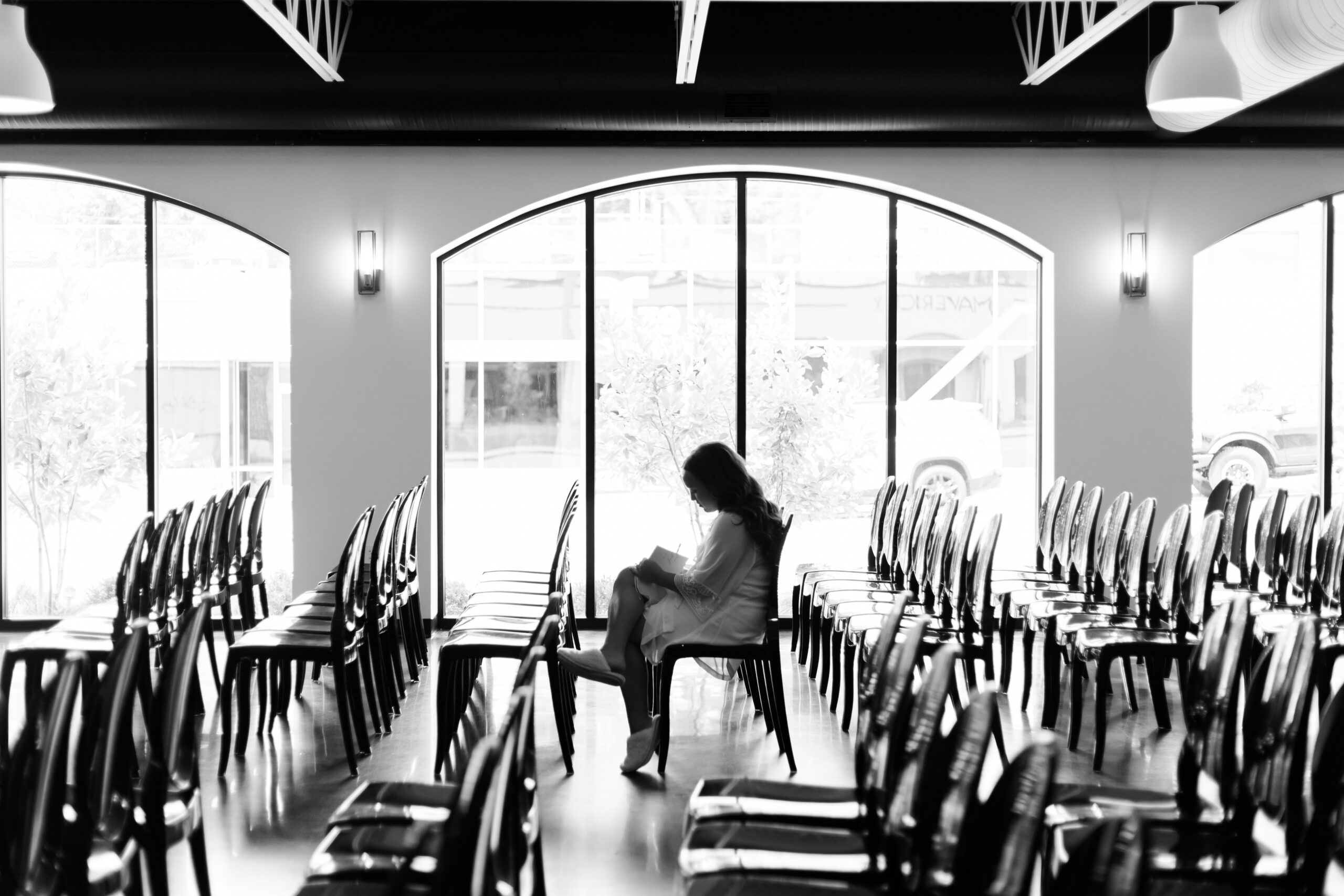 Bride sitting alone among rows of black chairs in a bright ceremony space, reading a small notebook before the wedding.