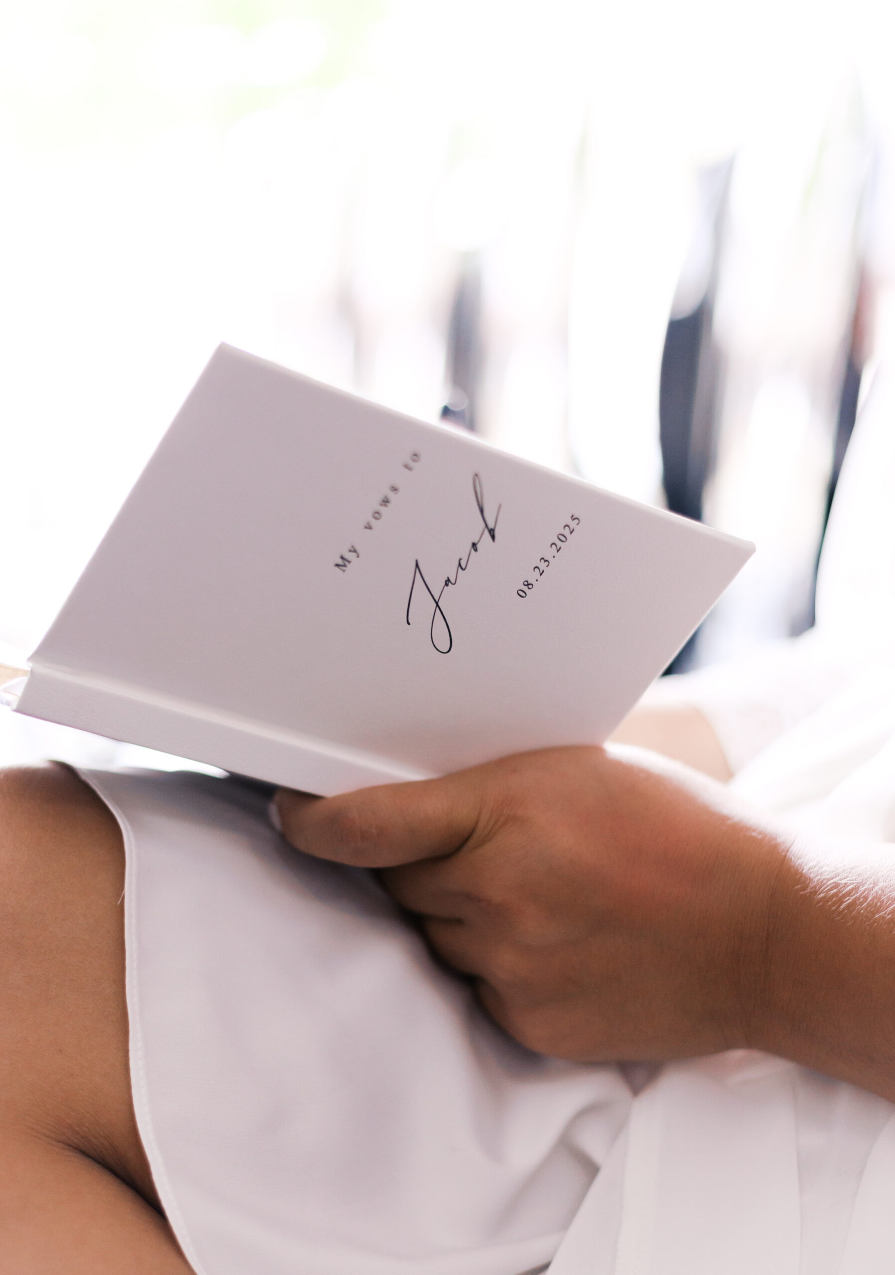Bride holding a personalized wedding vow book while seated in soft window light before the ceremony.