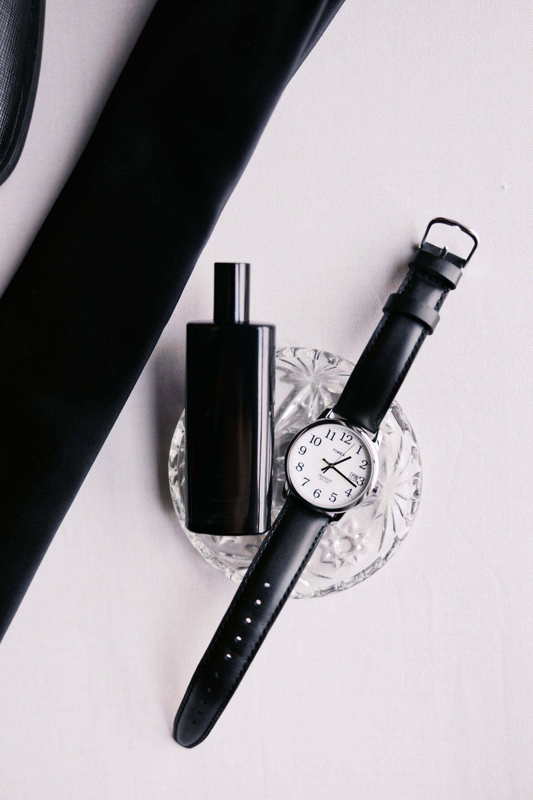 Groom’s accessories flat lay with black tie, cologne, and wristwatch styled on a neutral background.