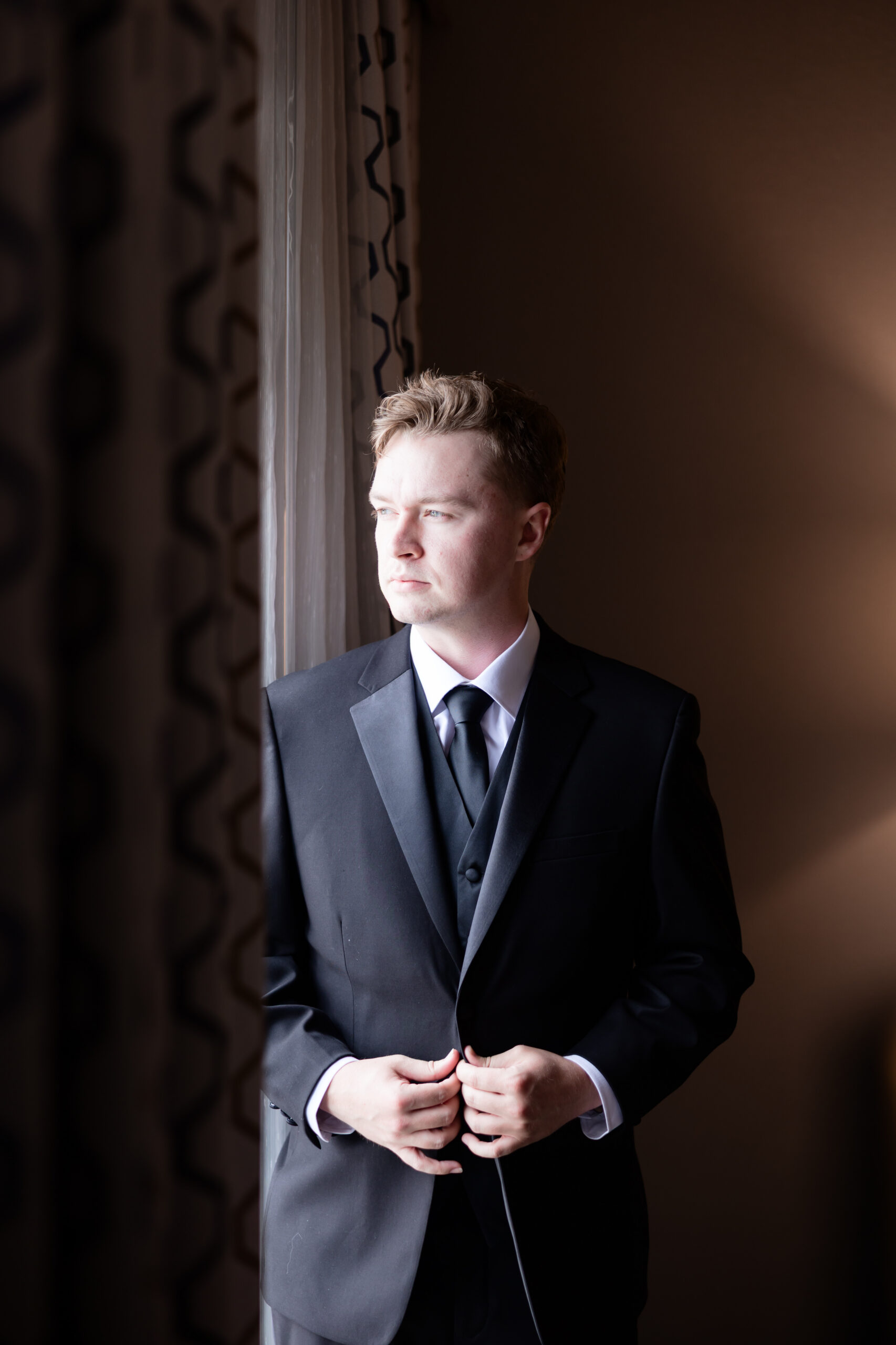 Groom in a black tuxedo stands by a window, adjusting his jacket in natural light before the ceremony.