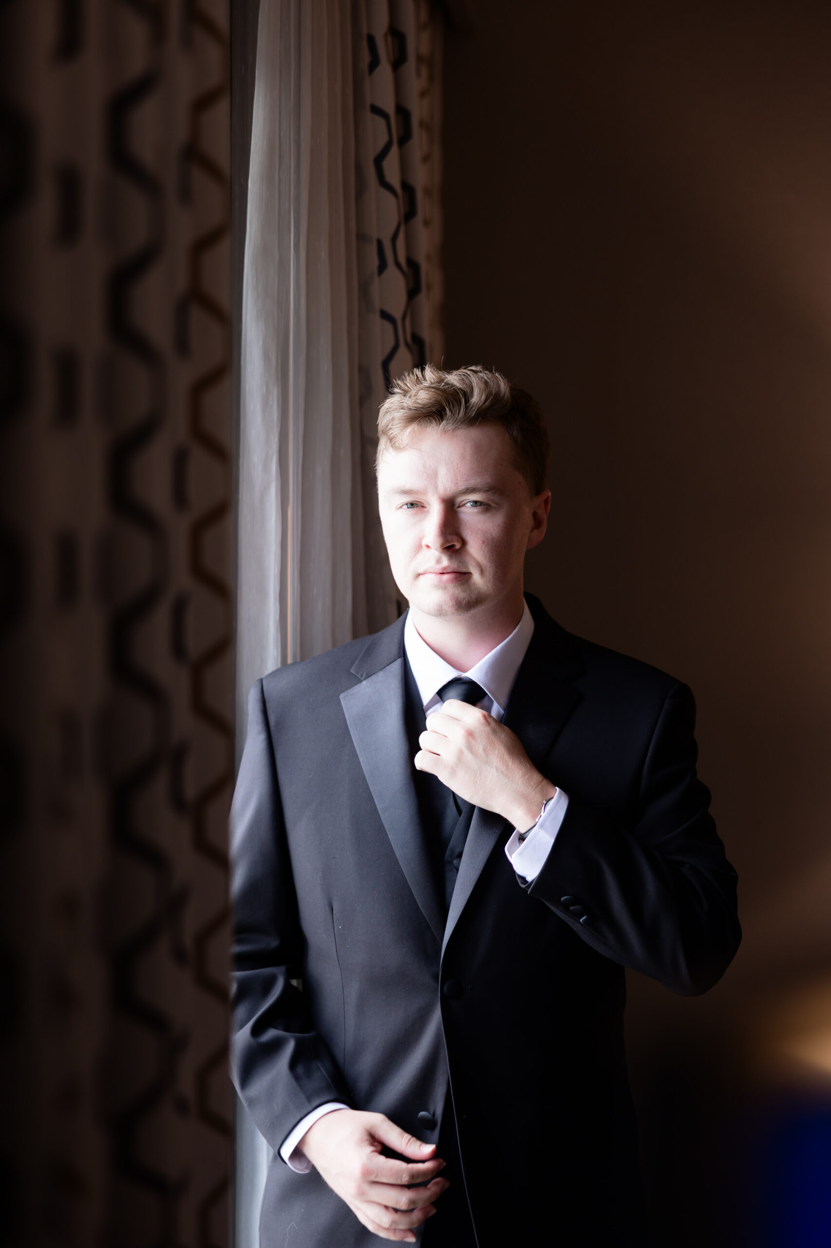 Groom adjusts his black tie while standing near a window during wedding preparations at The Maverick in Kansas City.