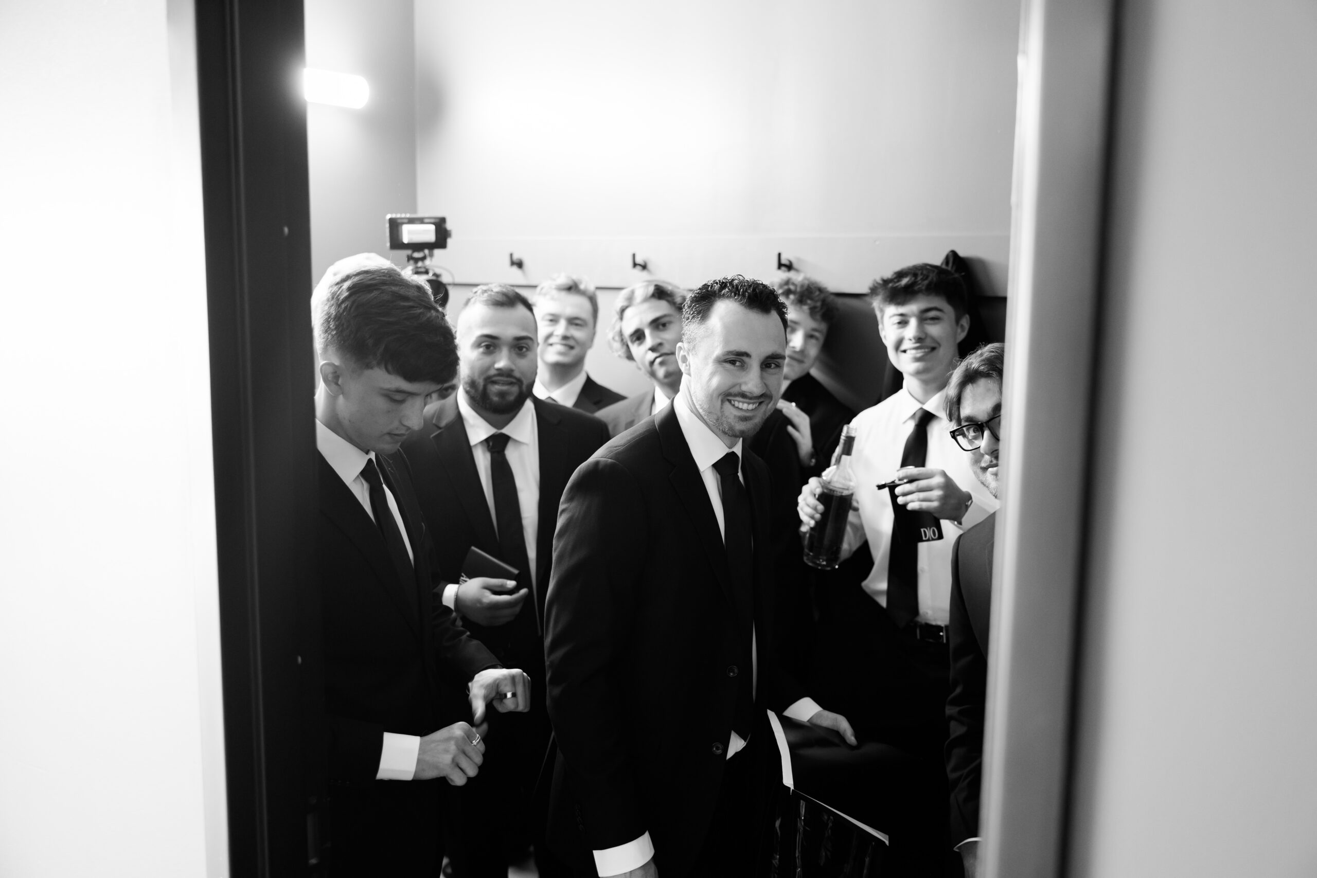 Black-and-white candid of groomsmen gathered in a hallway, dressed in black suits before the ceremony.