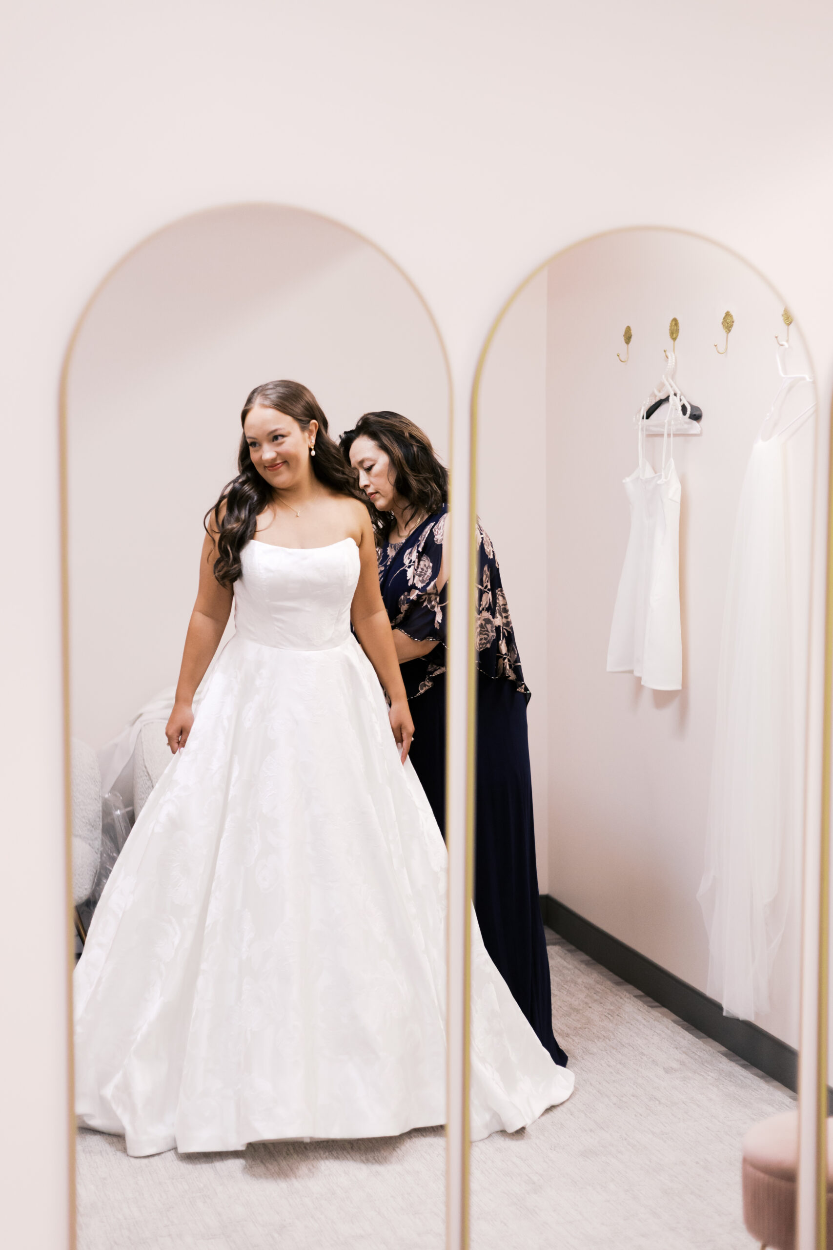 Bride stands in front of a mirror as a family member helps fasten her wedding dress in a bright dressing room.