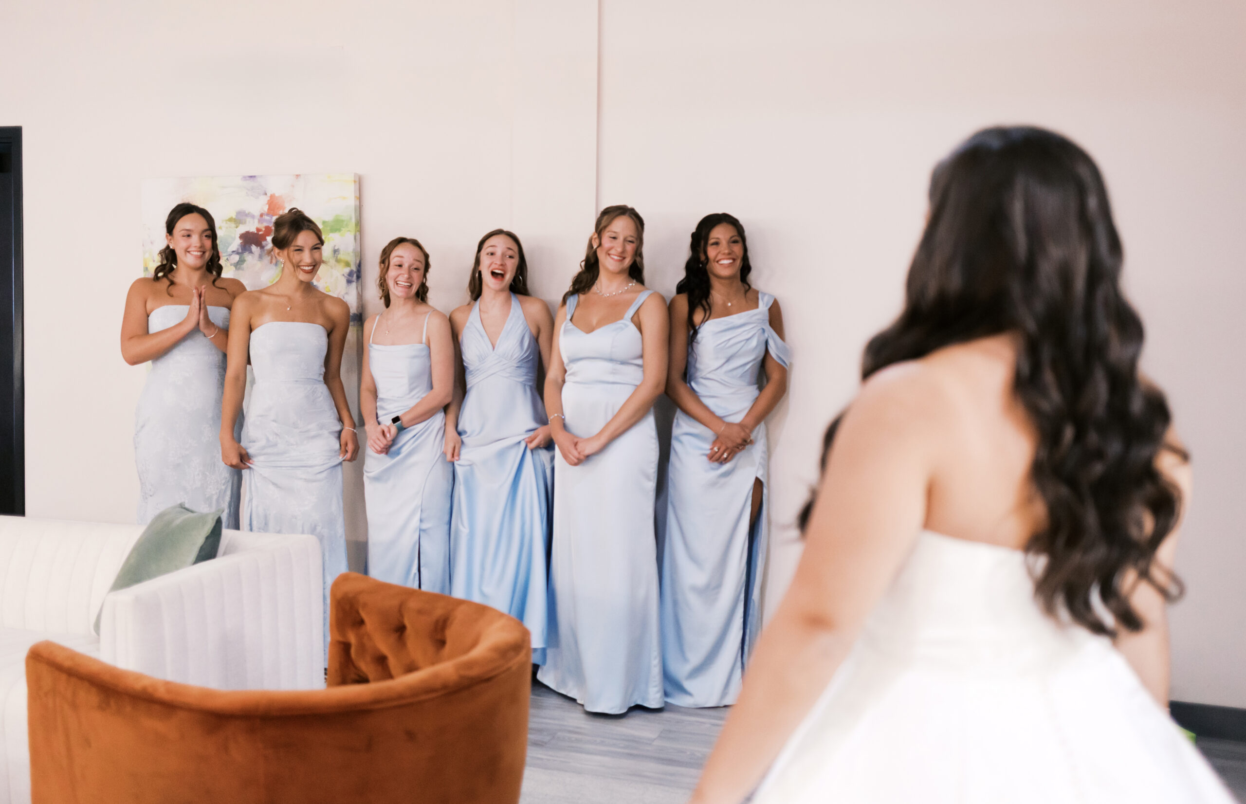 Bride enters the room as bridesmaids in light blue dresses react together during a first-look moment.