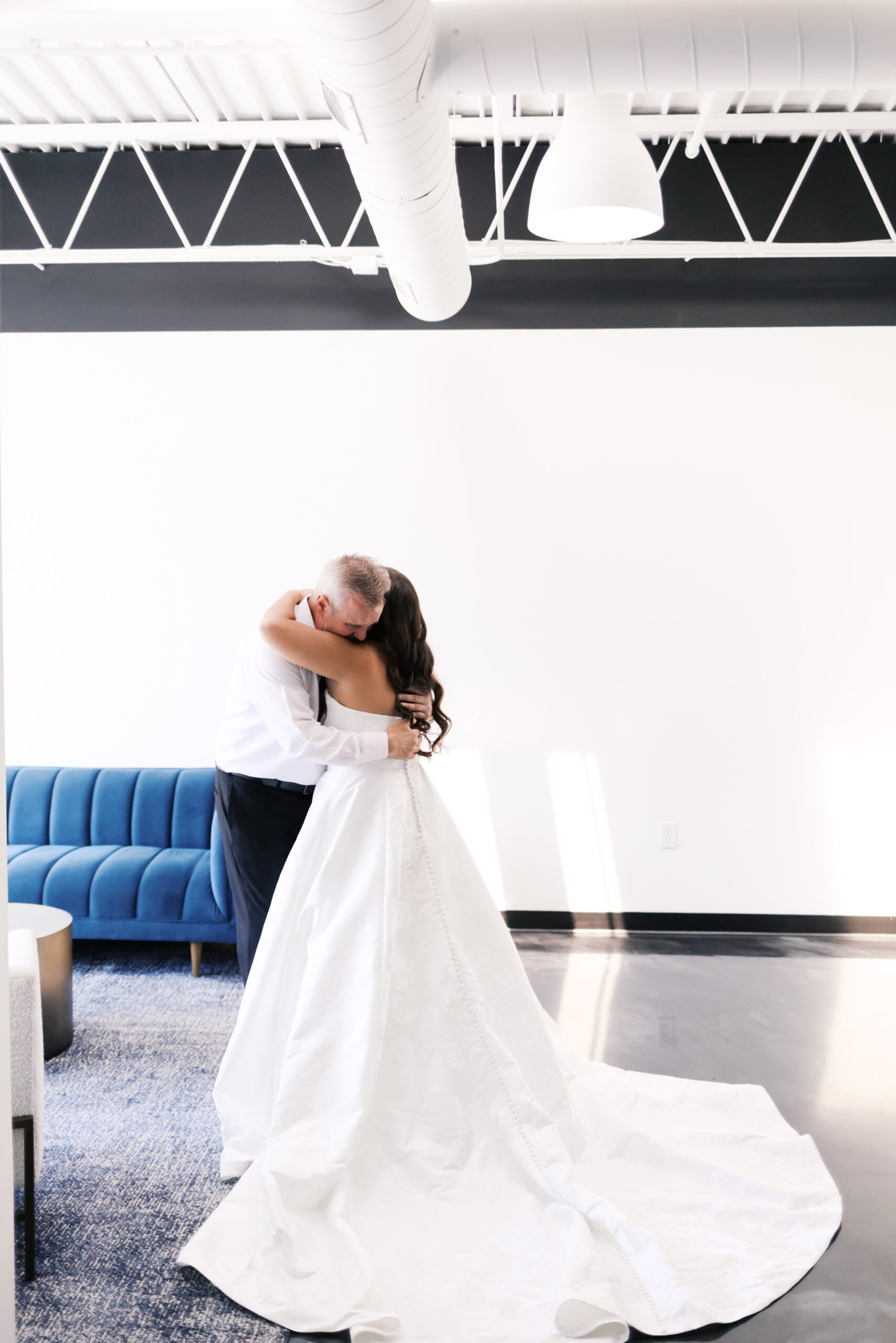 Bride and her father embrace in a bright room, with the wedding dress train flowing across the floor.
