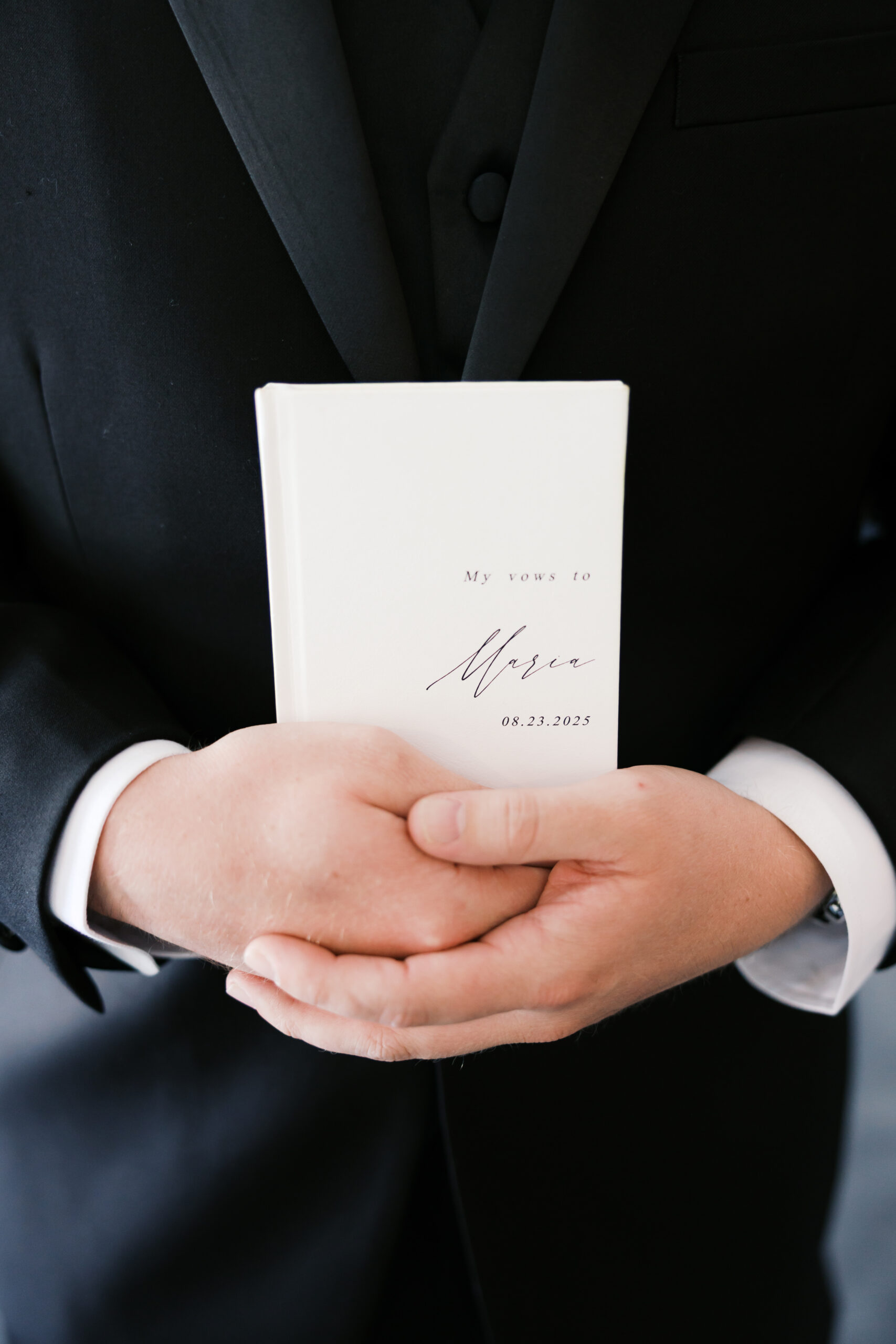 Groom holds a personalized wedding vow book against a black tuxedo, showing names and wedding date.