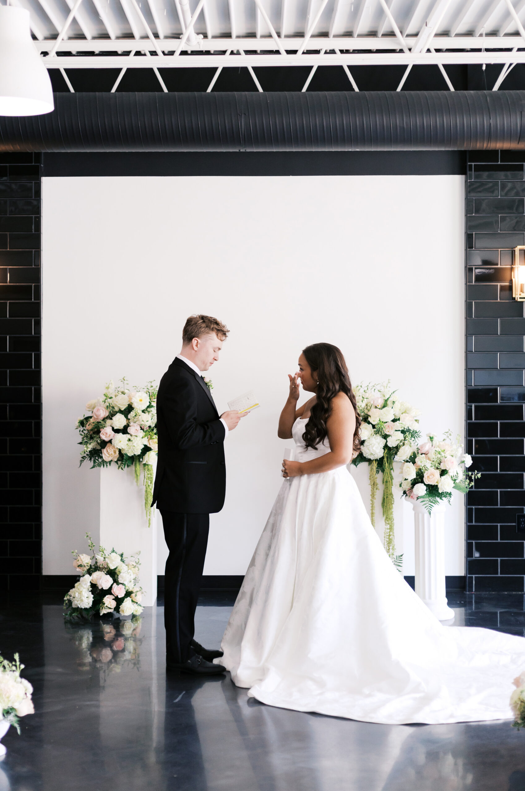 Bride and groom standing across from each other, groom reading vows from a small book as bride wipes away a tear.