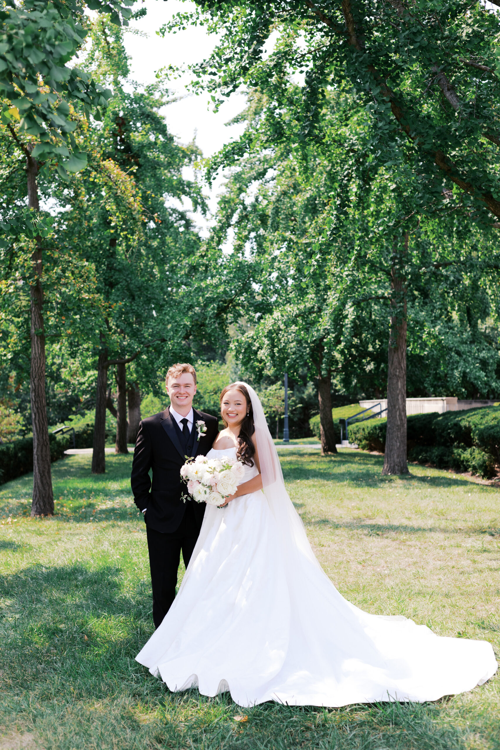 Outdoor portrait of the bride and groom smiling at the camera as they stand in the grass amongst tall trees.